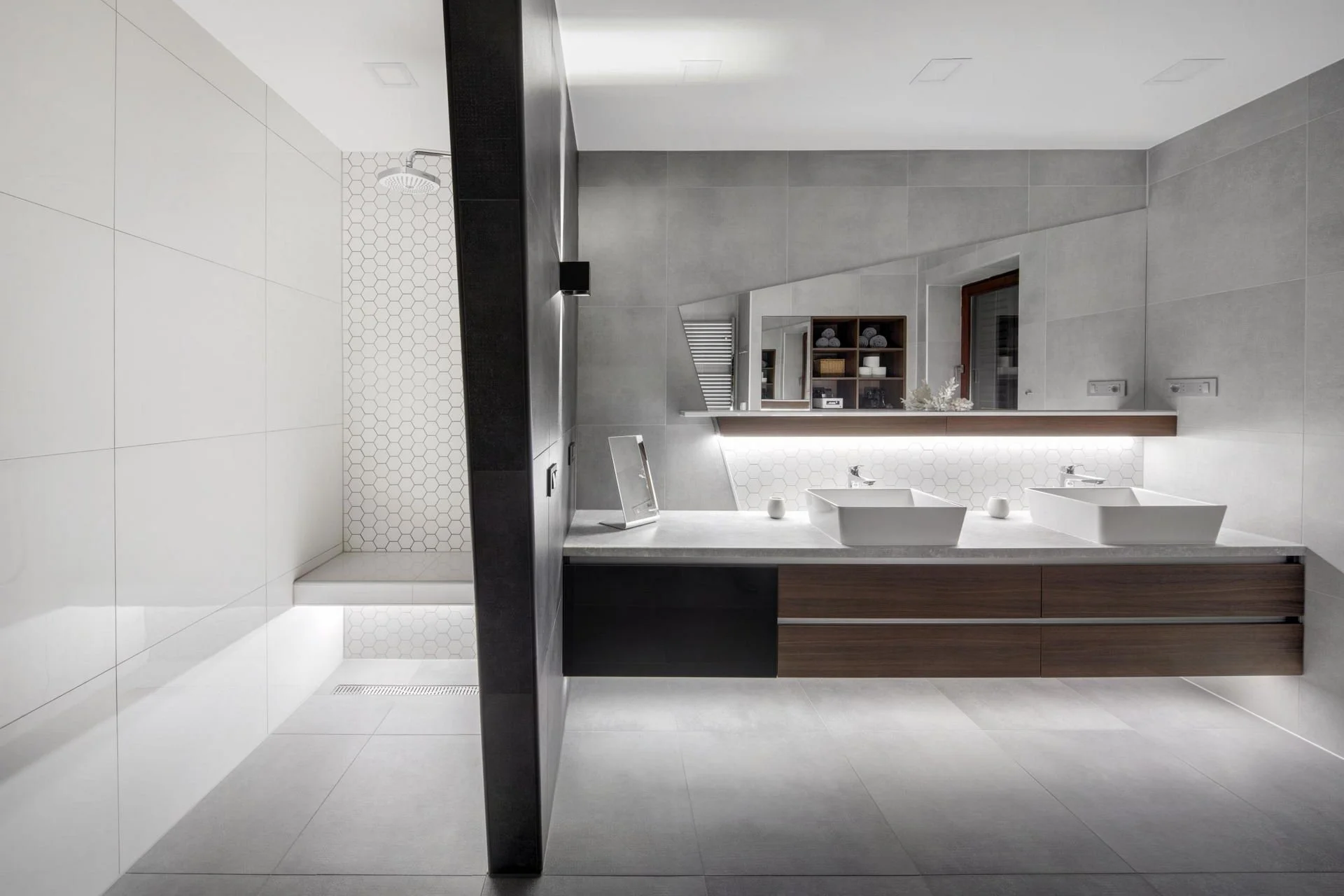 Modern bathroom with double sinks, gray tiles, large mirror, and adjacent shower area with white hexagonal tiles.