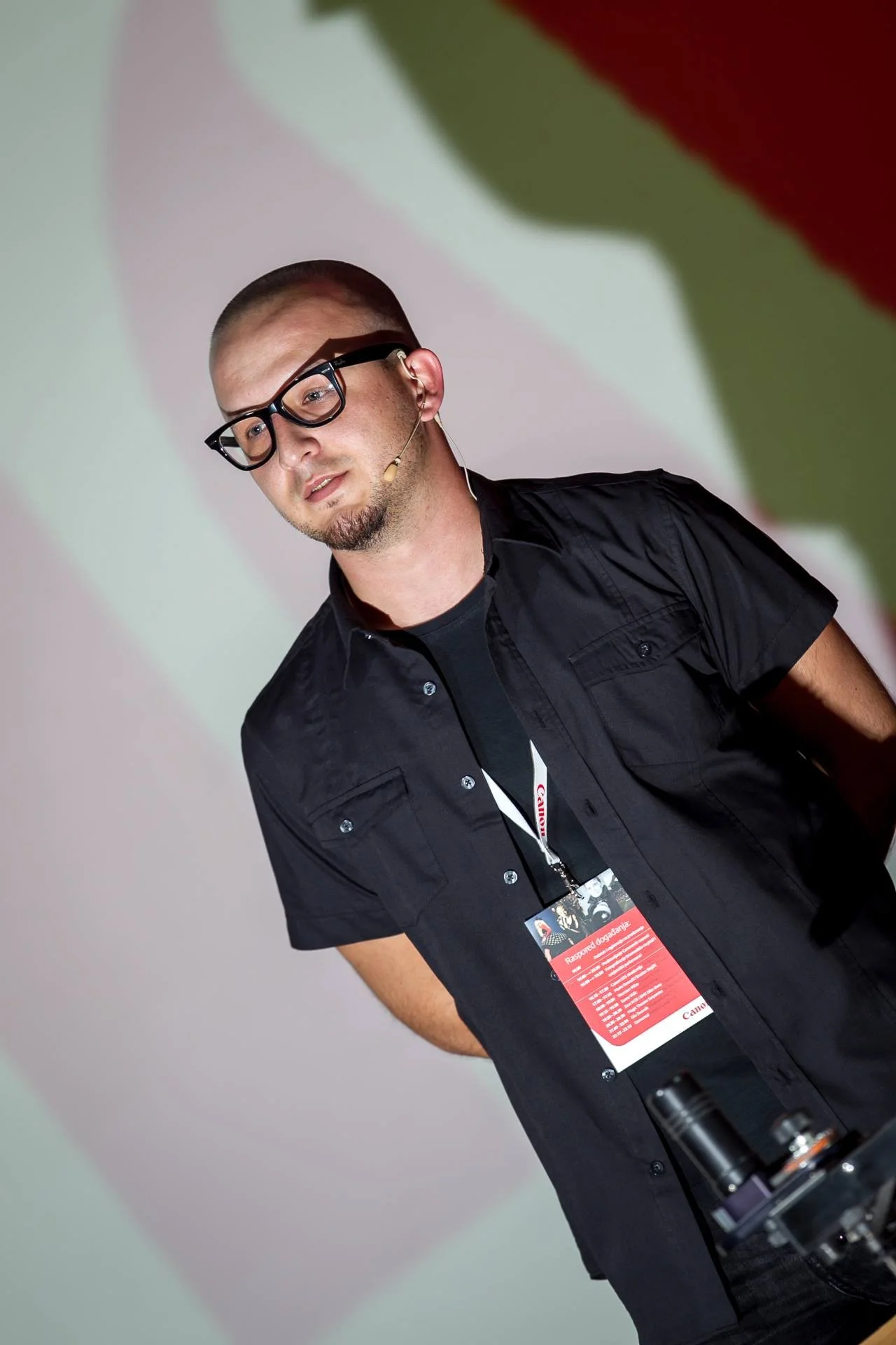 A young man with a buzz cut, glasses, and a beard speaking at a conference or presentation, wearing a black shirt, with a microphone headset, and a conference badge around his neck.