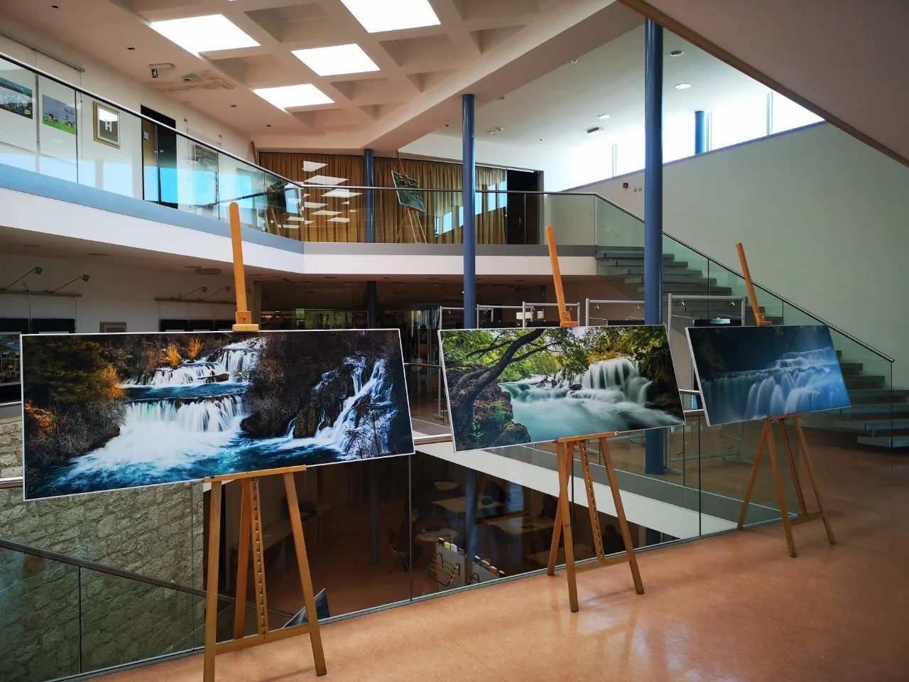 Gallery displays three landscape photographs of waterfalls and rivers on easels inside a modern building with glass railings and staircases.