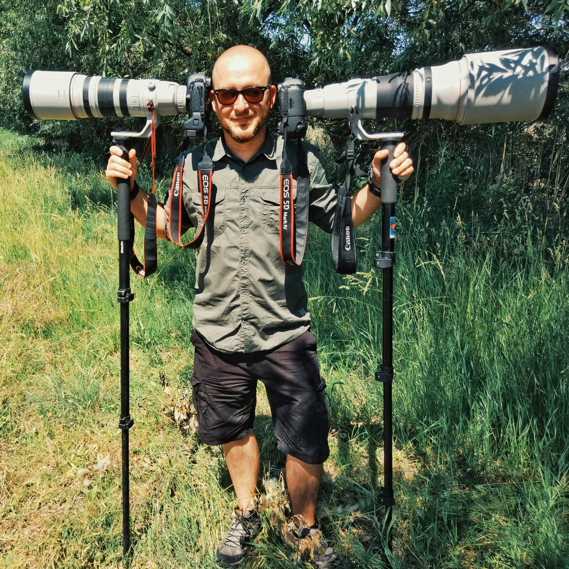 A man with sunglasses, a gray shirt, and black shorts standing in a grassy field holding a large camera lens with a tripod mount over his shoulders.