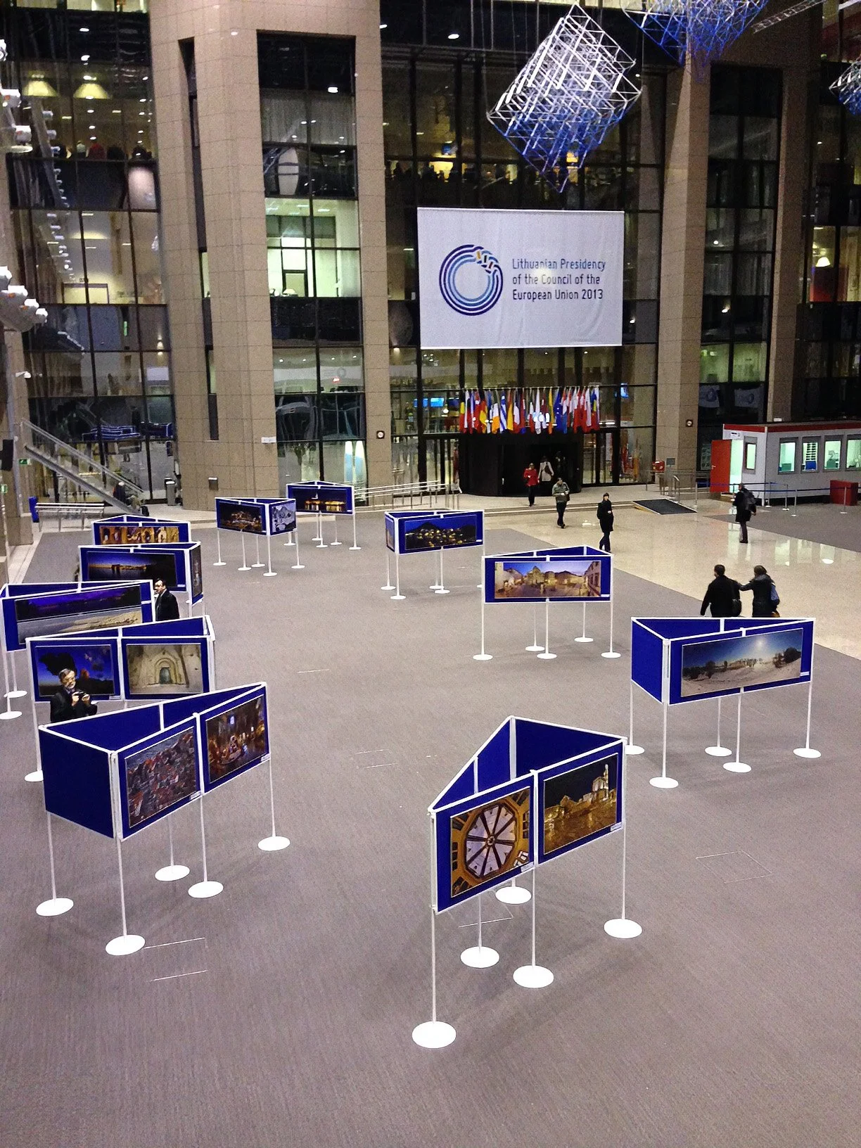 Exhibition hall with multiple display panels showing photographs, a large banner on the wall reads 'Lithuanian Presidency of the Council of the European Union 2013', and flags hang from the ceiling.