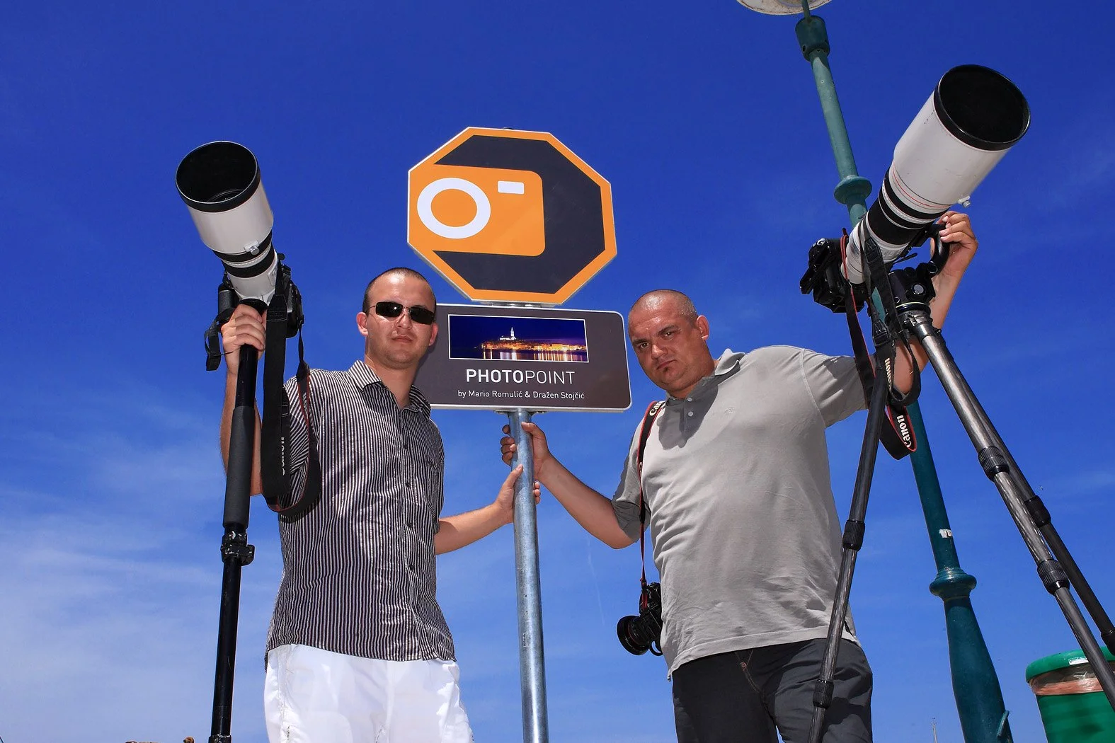 Two men standing under a clear blue sky, holding cameras with long telephoto lenses, near a sign that reads 'Photopoint' with a smaller sign underneath and a cityscape photo. One man is wearing sunglasses and a striped shirt, the other is in a light-