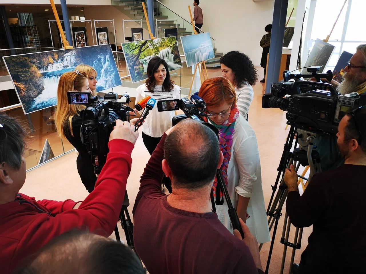 A group of people, including journalists with cameras, interviewing women in an art gallery showcasing landscape photographs of waterfalls and nature scenes.
