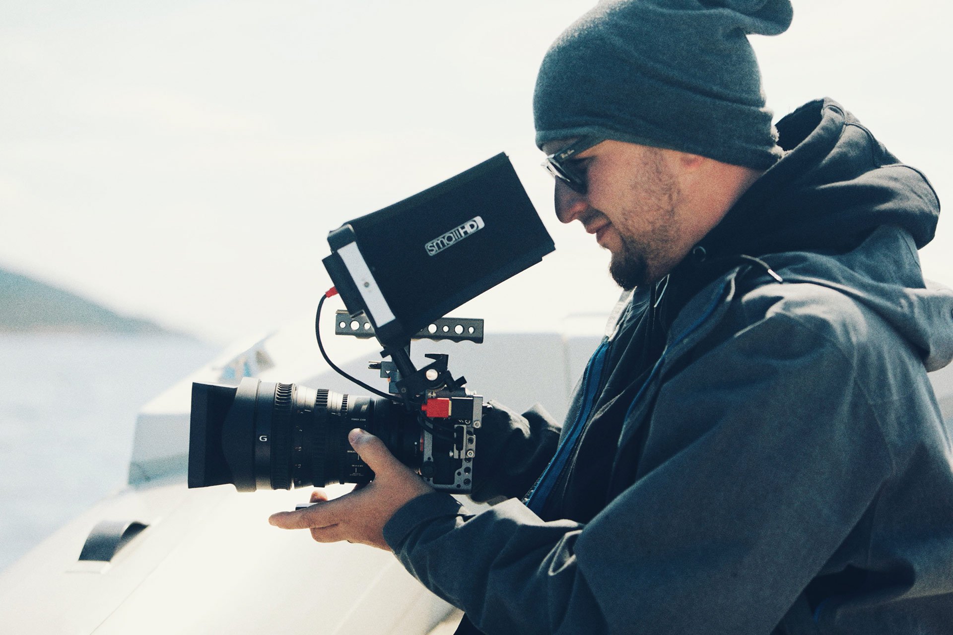 A man wearing a dark hoodie, sunglasses, and a beanie hat operating a professional camera mounted on a stabilizer, with a bright background suggesting an outdoor setting.