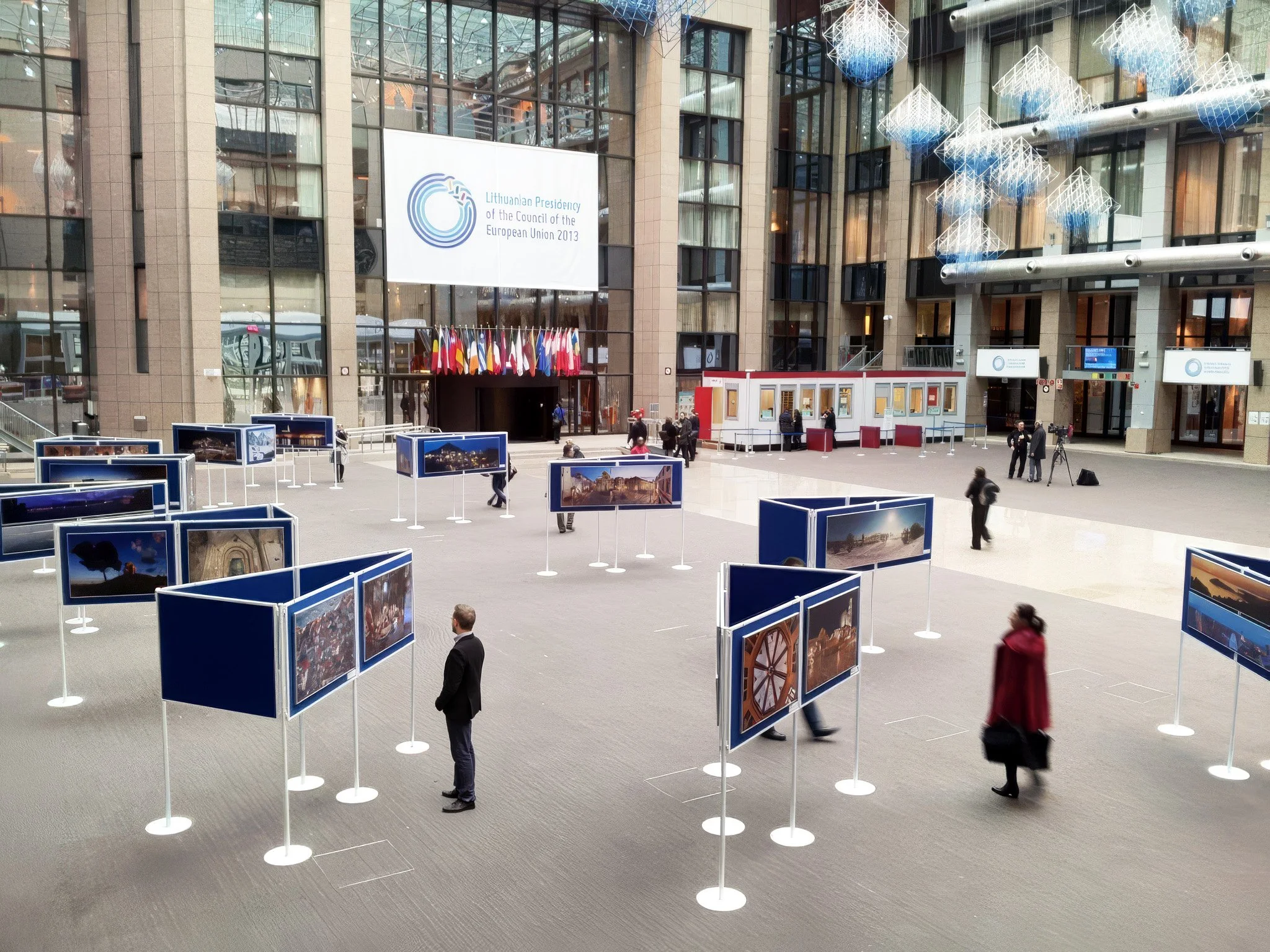 An indoor exhibition space with framed photographs arranged on blue display panels, in front of a large modern building with glass windows. Several people are viewing the photographs, and a banner displayed in the background reads 'Lithuanian Preside