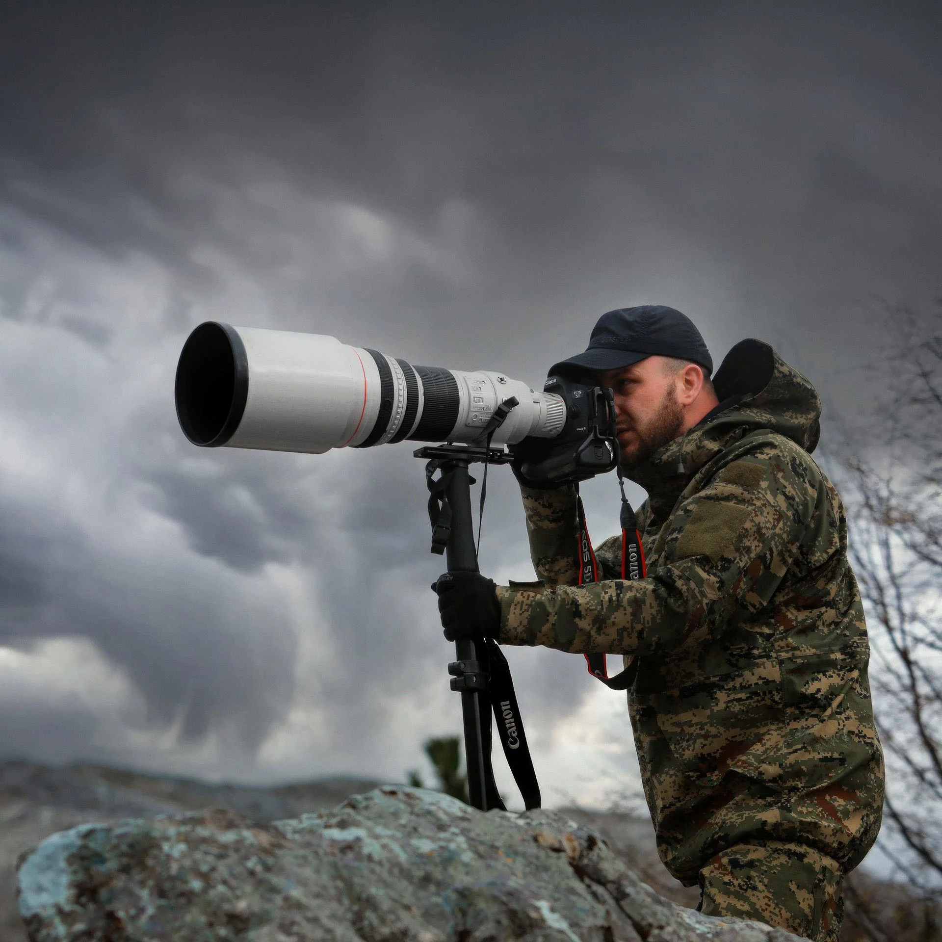 A man in camouflage clothing and a black cap aims a large telephoto camera lens on a tripod, outdoors on a cloudy day.
