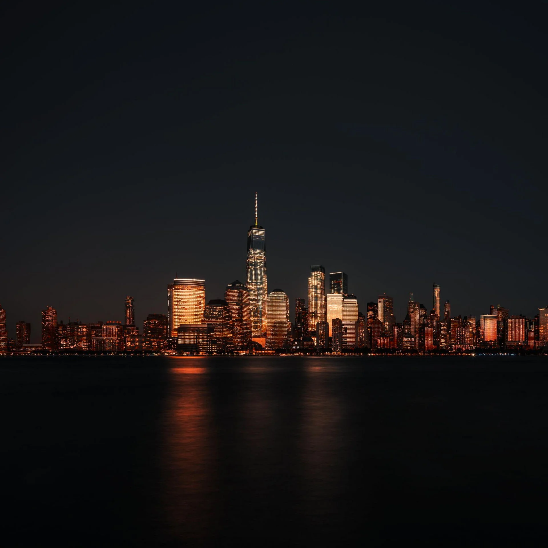 Nighttime city skyline of Manhattan with illuminated buildings, including the One World Trade Center, reflected on the water.