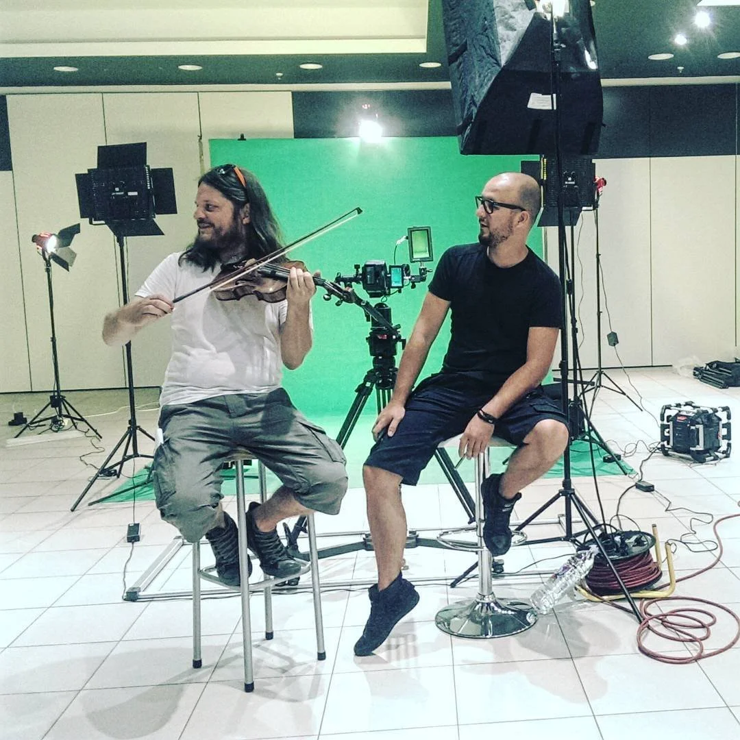 Two men on a set with a green screen, surrounded by lighting and camera equipment. One man is playing a violin while the other sits on a stool, watching.