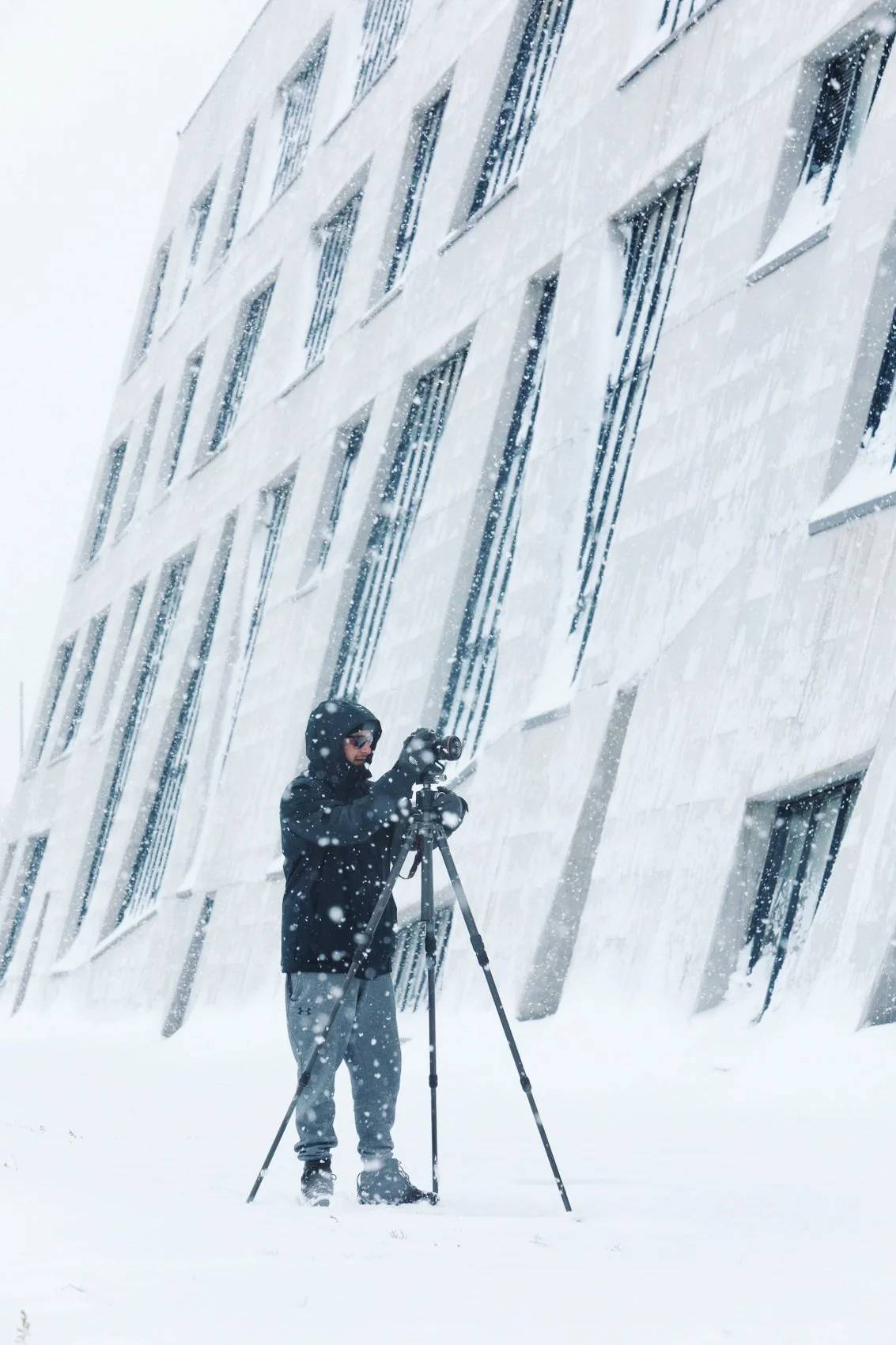 A person dressed in winter clothing, including a hood and gloves, is standing outside in a snowstorm, operating a camera on a tripod in front of a modern building with snow on its windows and wall.