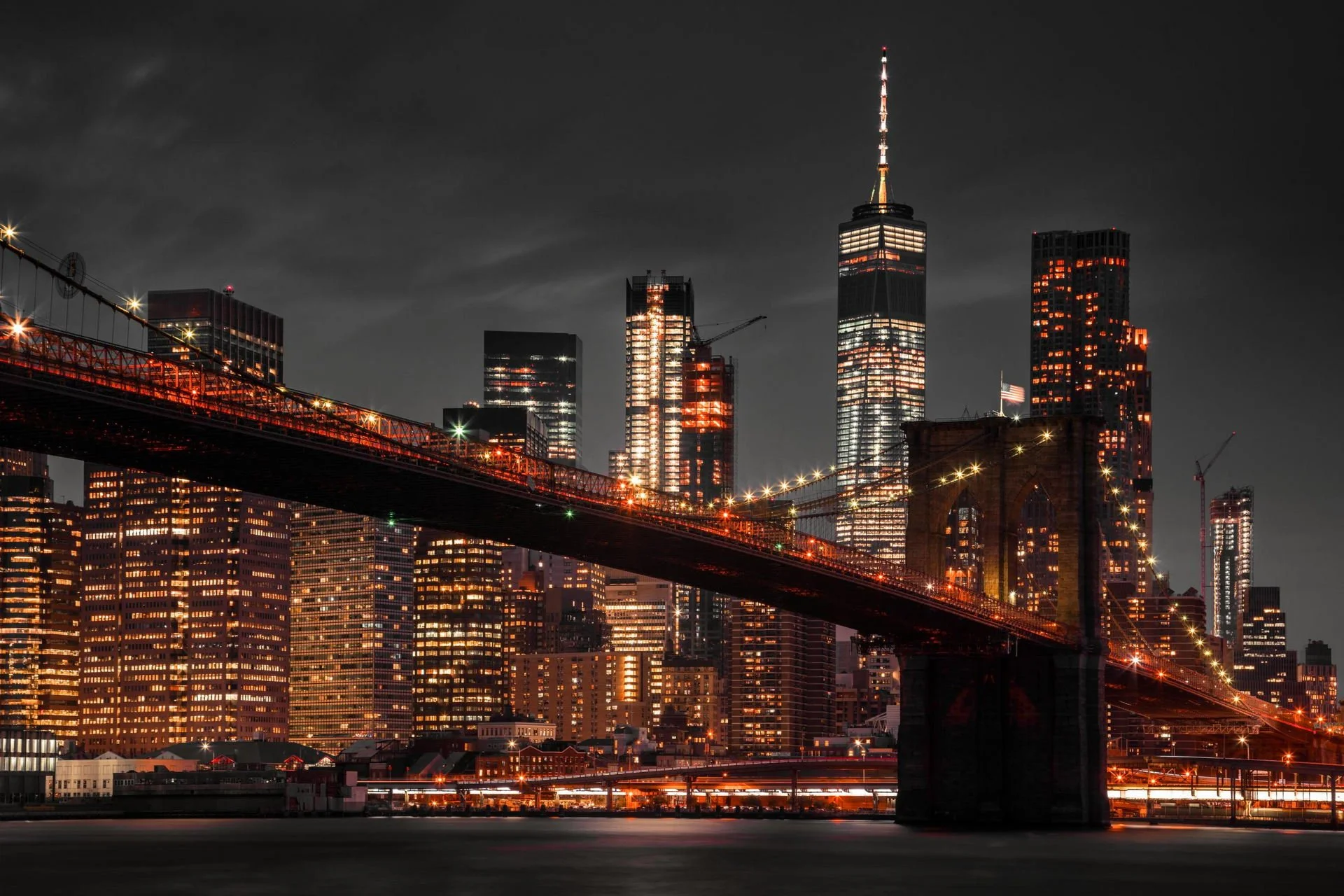 Nighttime view of the New York City skyline with illuminated skyscrapers and the Brooklyn Bridge in the foreground.