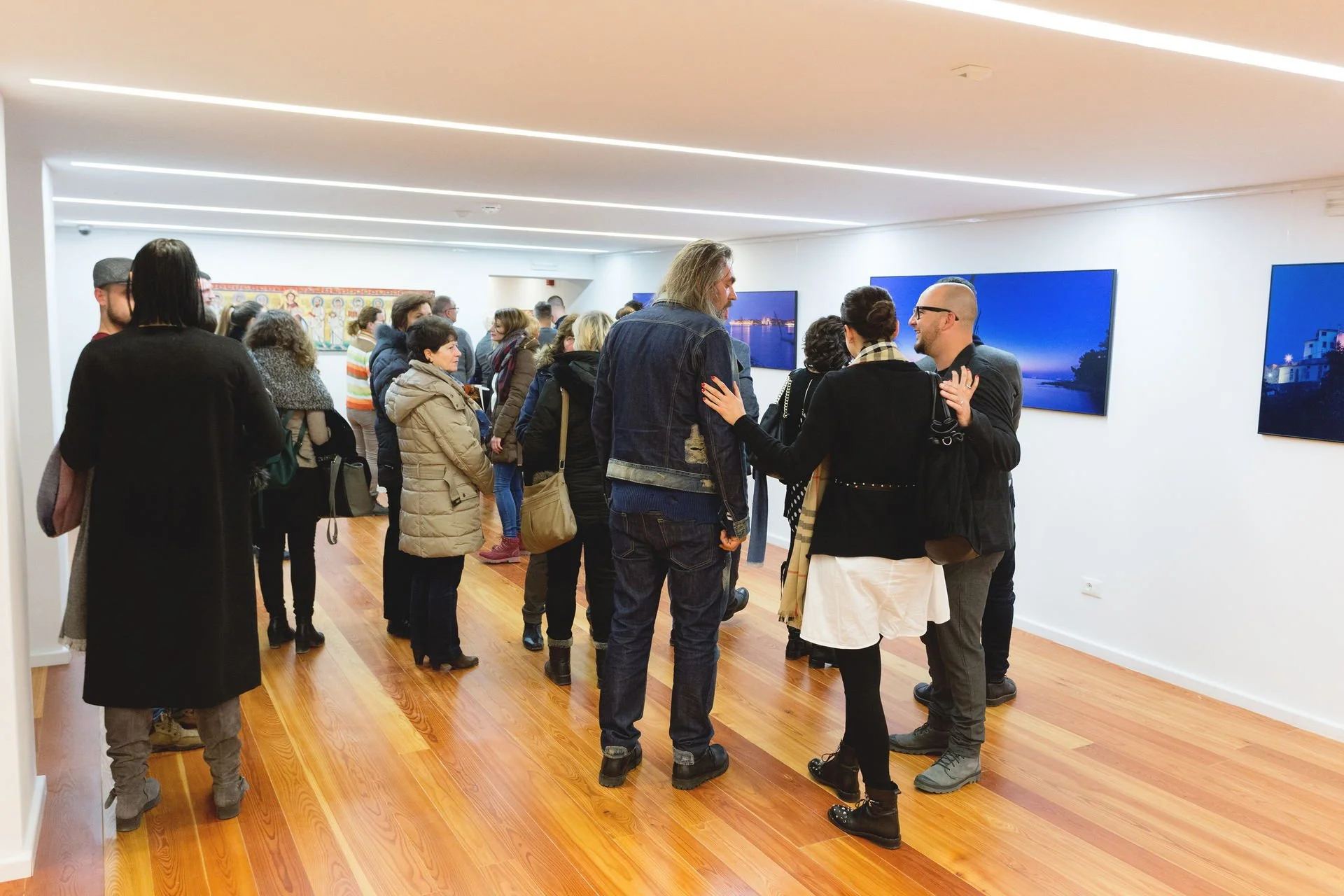 People viewing artwork in an art gallery, some engaging in conversation, with landscape photos displayed on the walls.