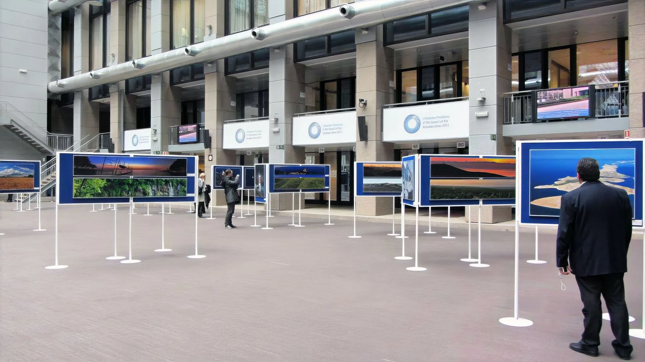 An outdoor art exhibit displaying landscape photographs on blue panels outside a modern building with glass windows and white signs.
