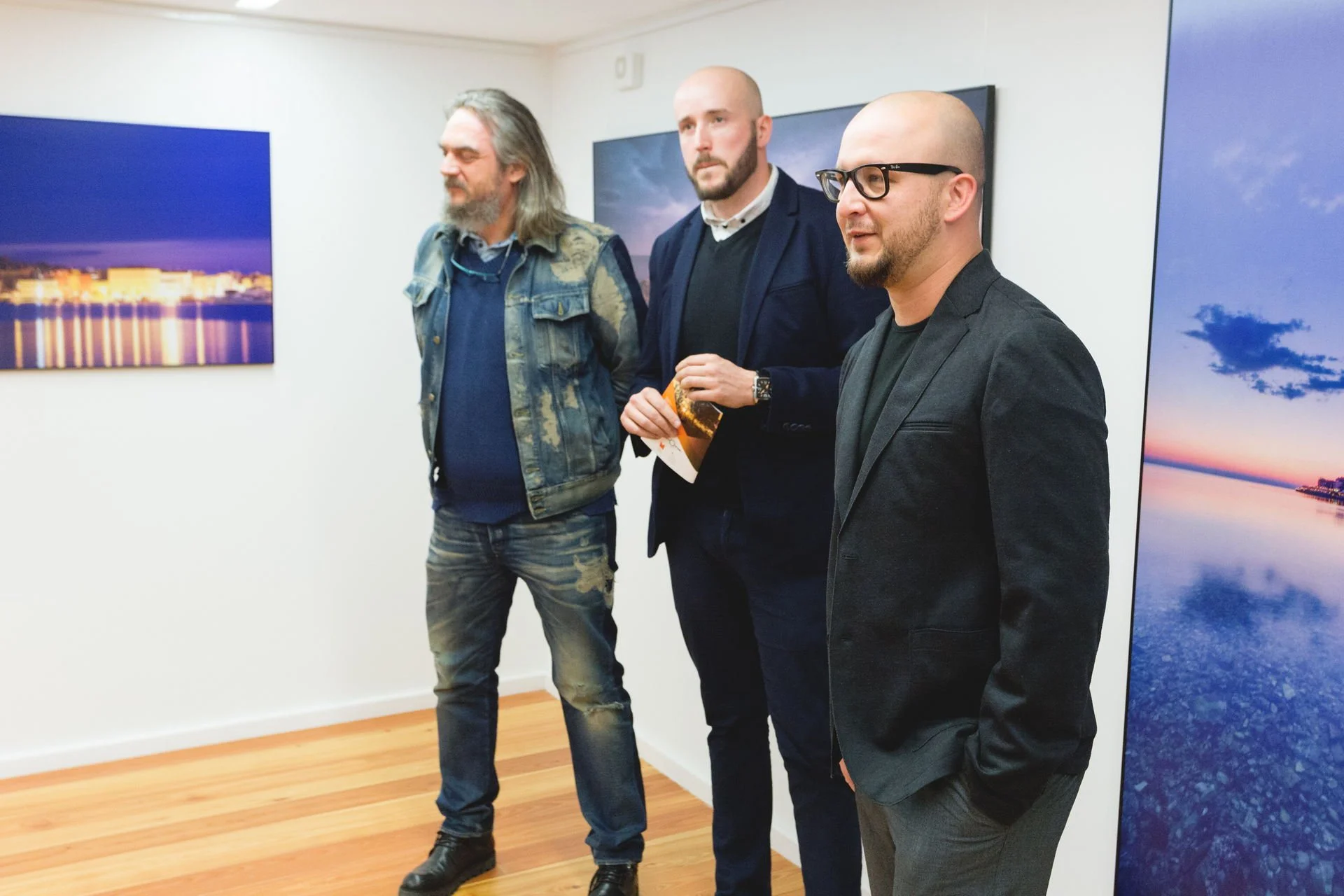 Three men standing in an art gallery, observing landscape photographs on the wall.