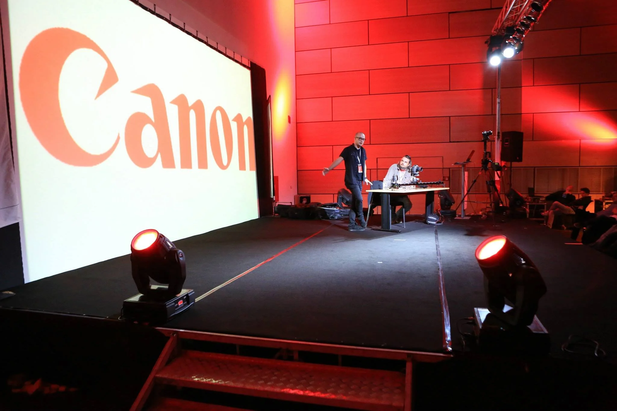 A stage at a conference or event with a large Canon logo projected on a screen. There are two men on the stage, one standing and one sitting at a table with audio or DJ equipment, with red stage lighting and seating for an audience.