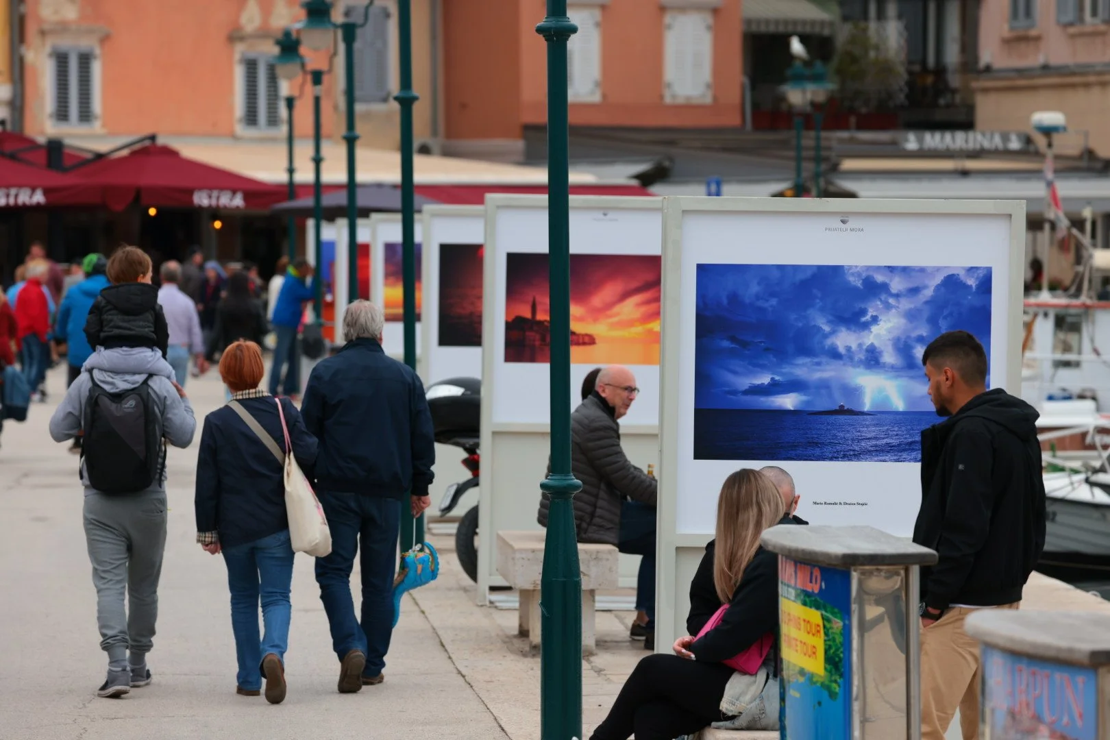 People walking along a waterfront with artwork displays and boats in the background.