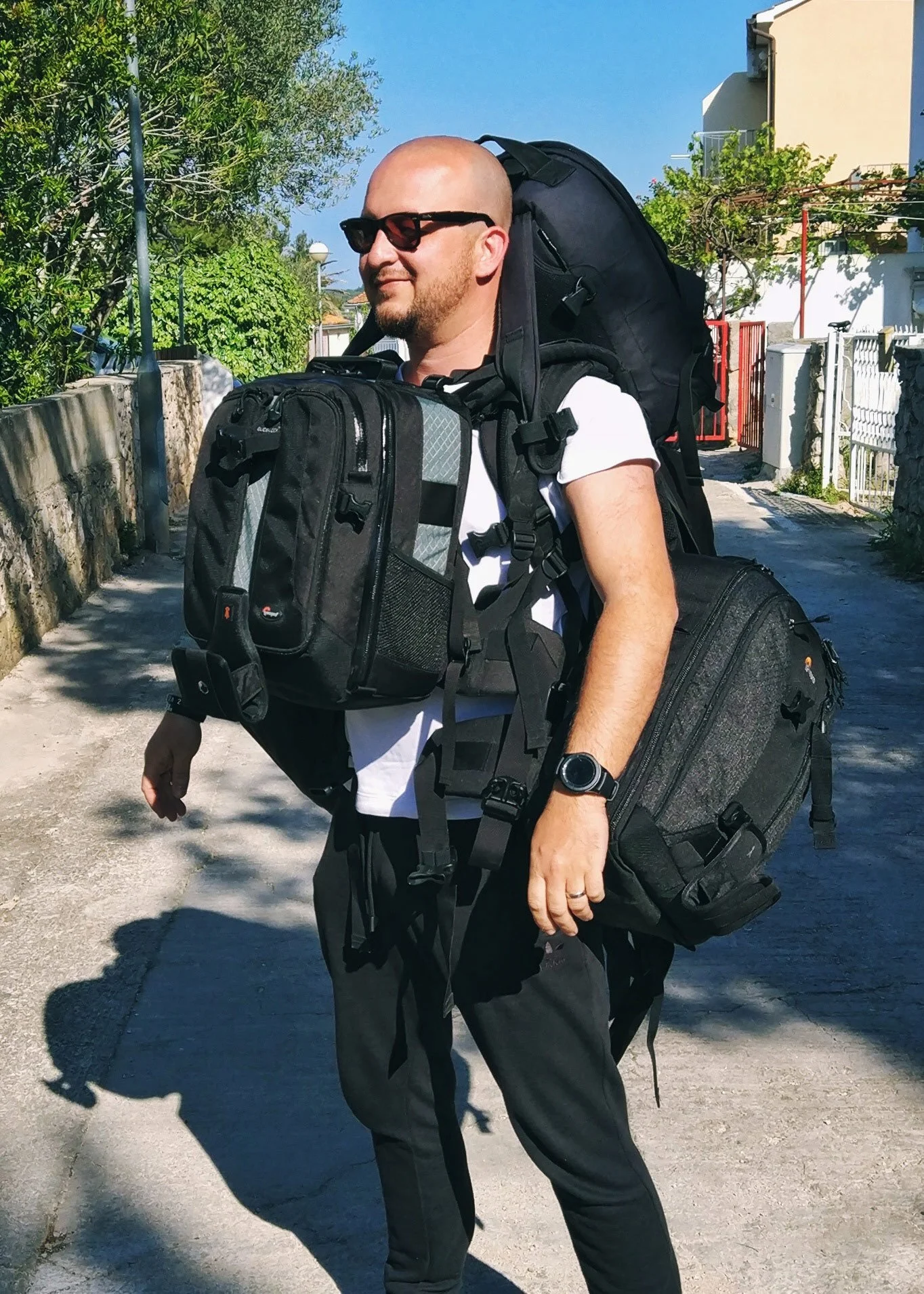 A man wearing sunglasses, a white t-shirt, and a black wristwatch standing outdoors with a large backpack and a smaller bag, on a sunny day with trees and houses in the background.