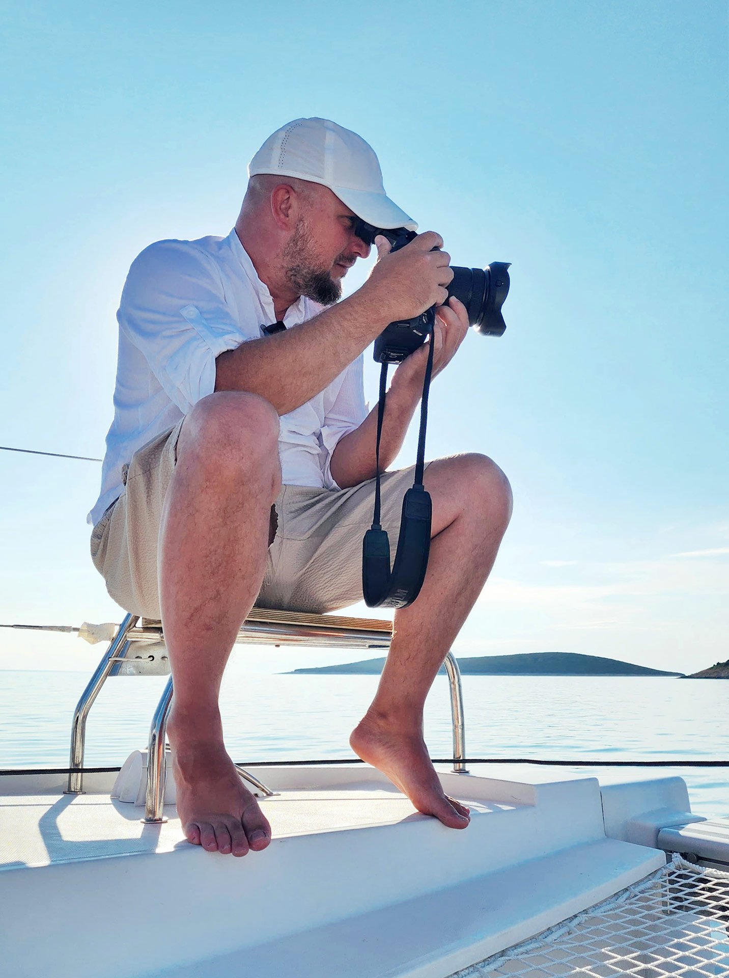 A man taking a photograph with a camera while sitting on the edge of a boat, with a calm sea and hilly islands in the background.