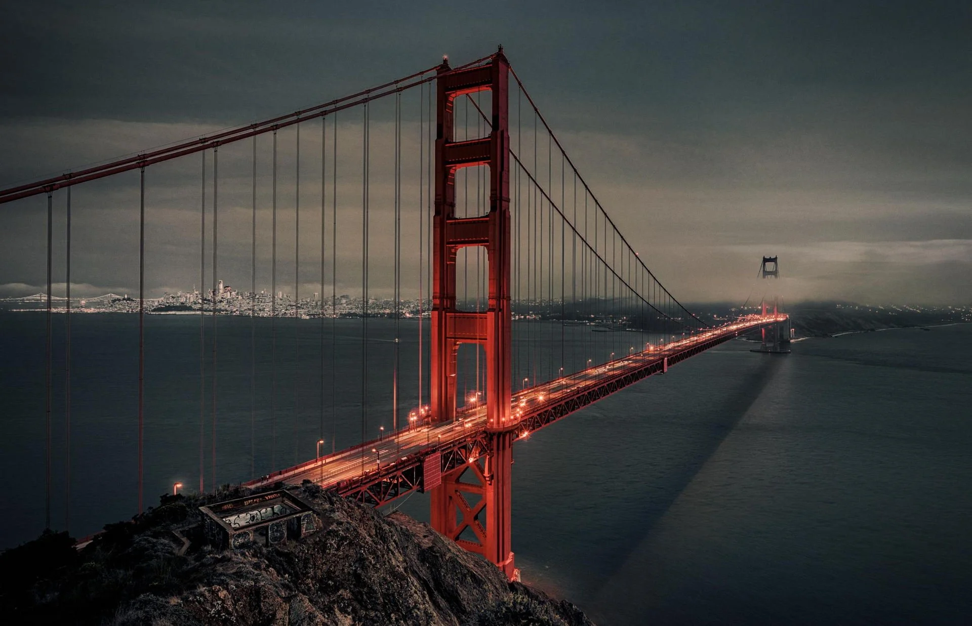 Night view of the Golden Gate Bridge in San Francisco, with city skyline in background and fog surrounding the bridge towers.