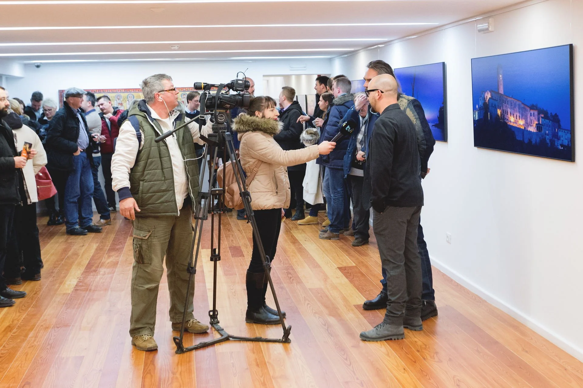 A woman interviews a man in a gallery, surrounded by other people, with multiple screens displaying scenic images on the wall.