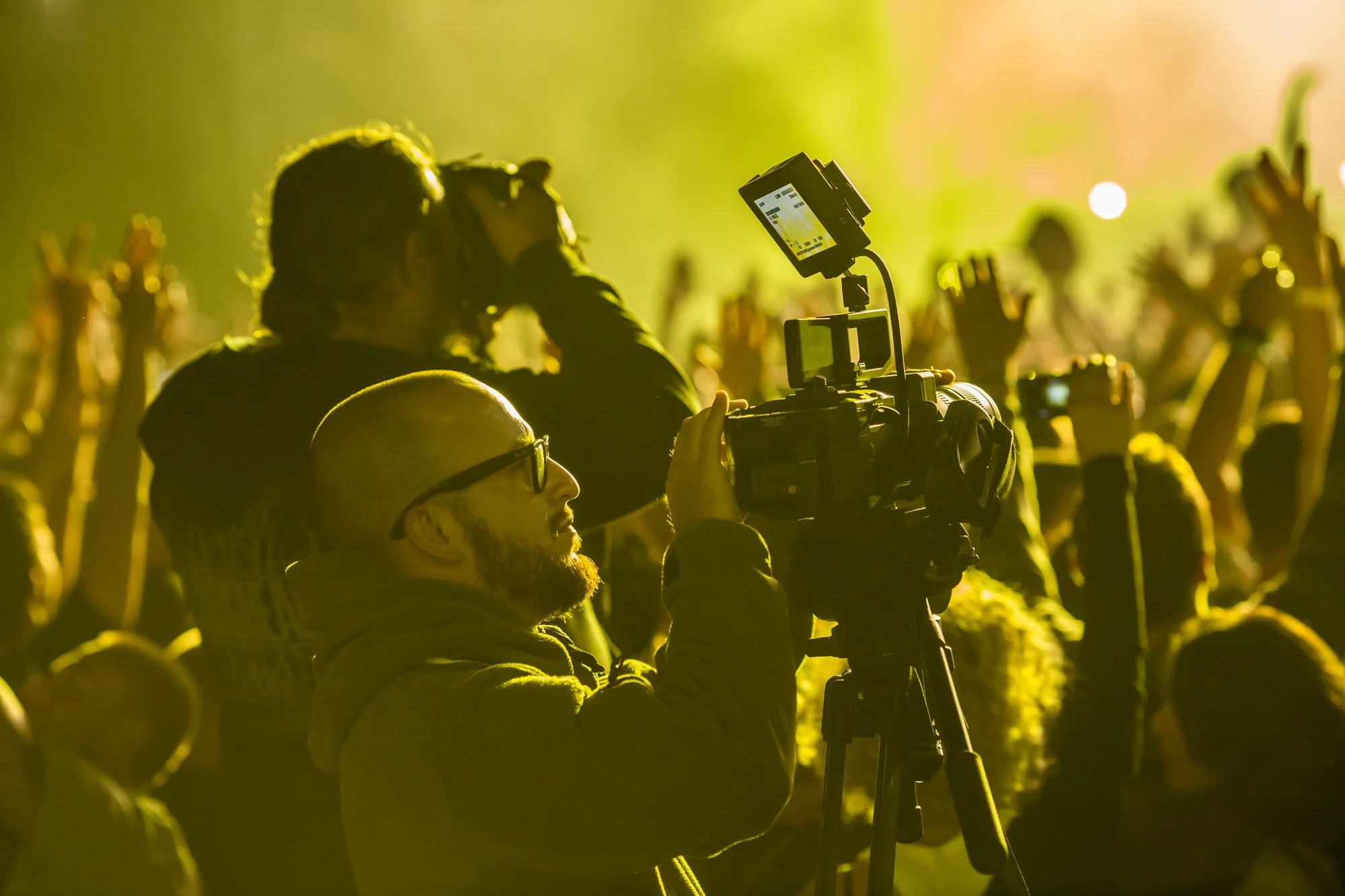 Dražen Stojčić, photographer from Osijek. A cameraman with glasses operating a camera at a lively event with many attendees and raised hands, illuminated by yellow lighting.