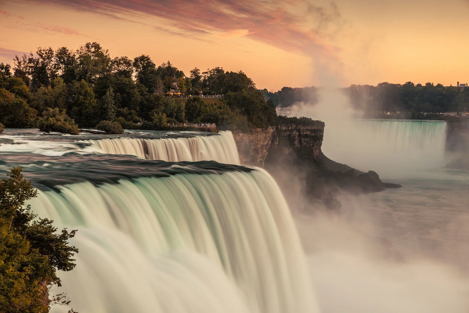 Niagara Falls at sunset with cascading waterfalls, mist rising, and a colorful sky.