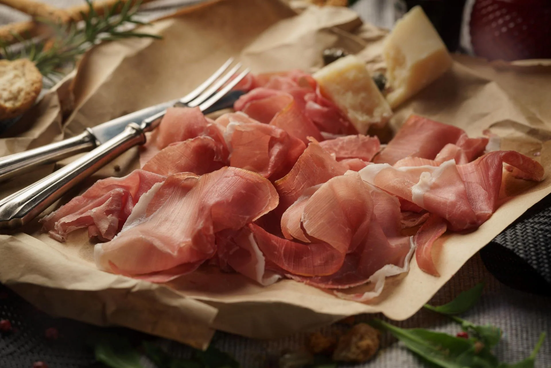 Assorted slices of prosciutto and other cheeses on parchment paper, with a fork and greenery in the background.
