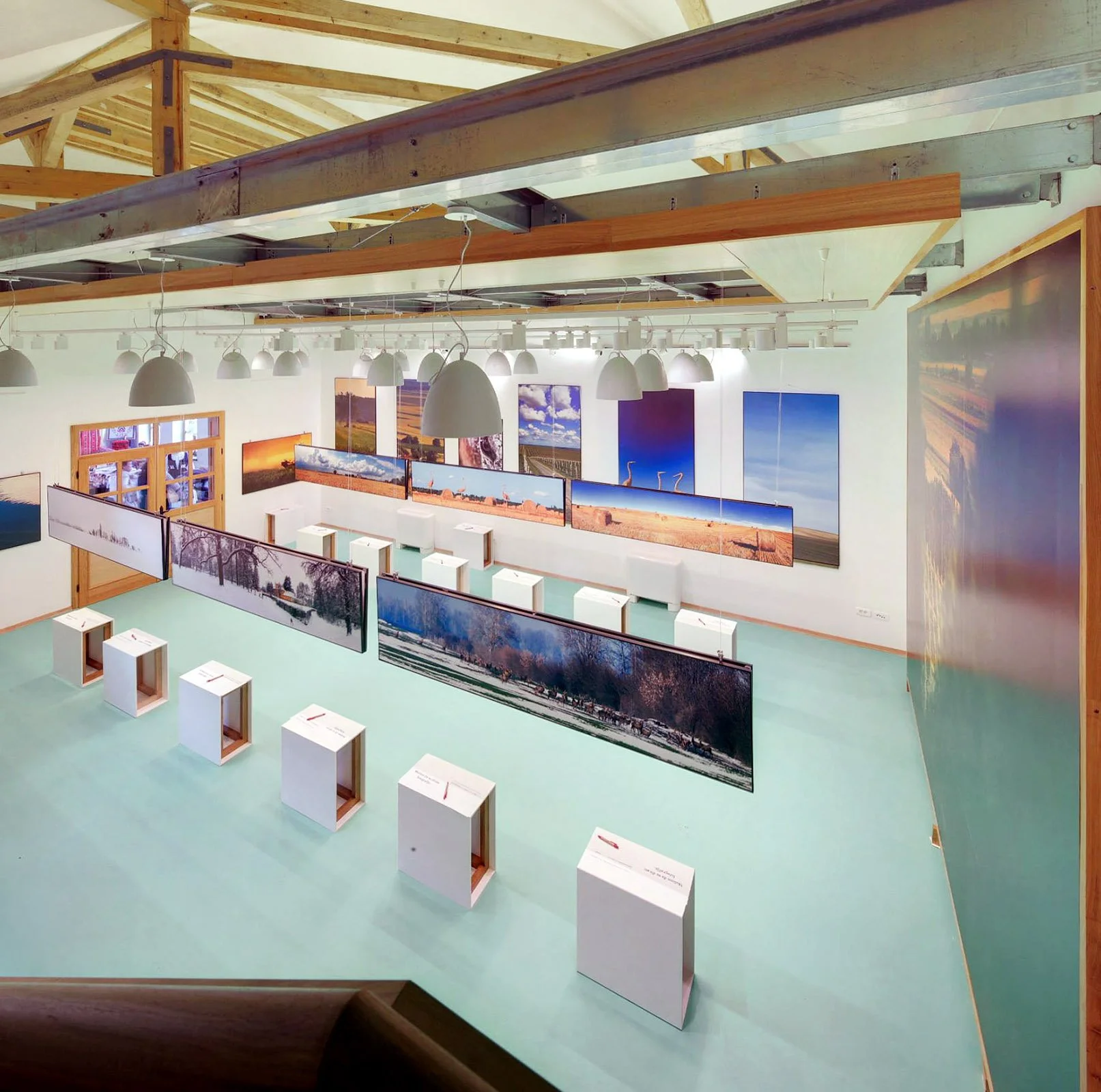 An art gallery with landscape photographs hanging on white walls, with paintings of skies, fields, and trees, and white pedestals on light green flooring.