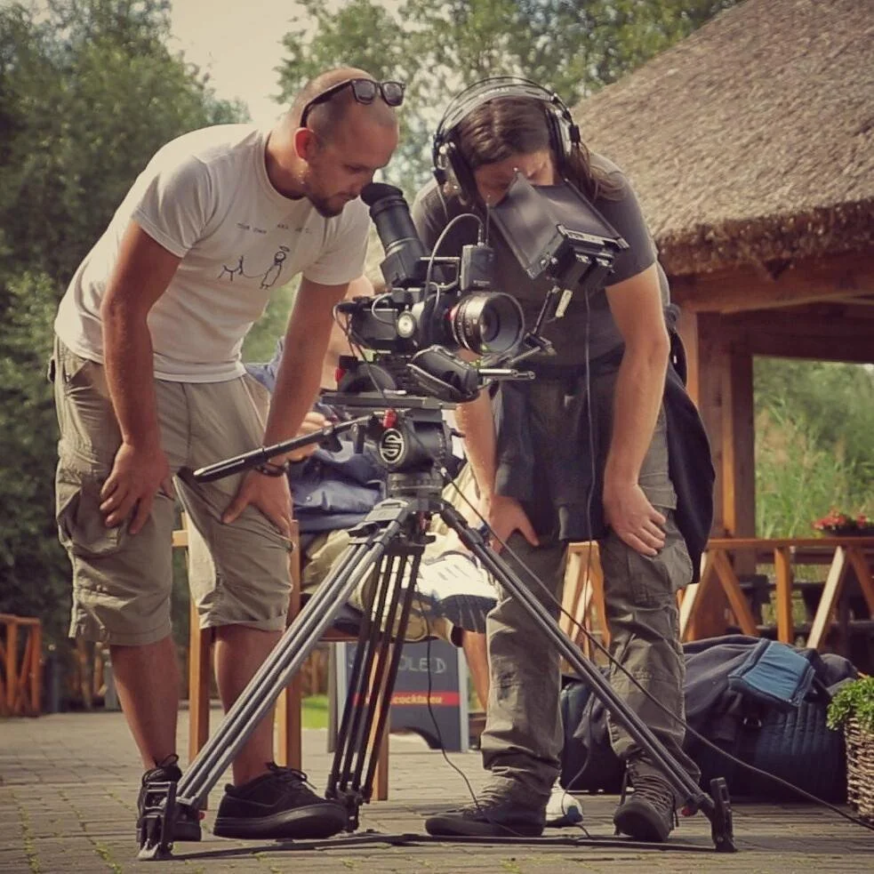 Two people working with a professional camera on a tripod outdoors near a wooden structure, with trees in the background.
