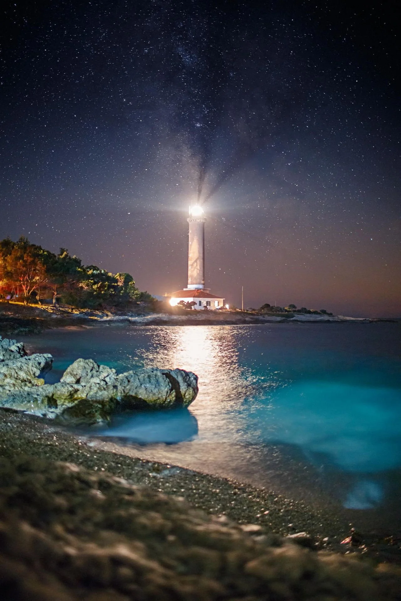 Dugi otok, Nighttime view of a lighthouse by the water with a starry sky and Milky Way overhead. The lighthouse's light is shining brightly, reflecting on the water. Rocks and some trees are visible in the foreground along the shore.