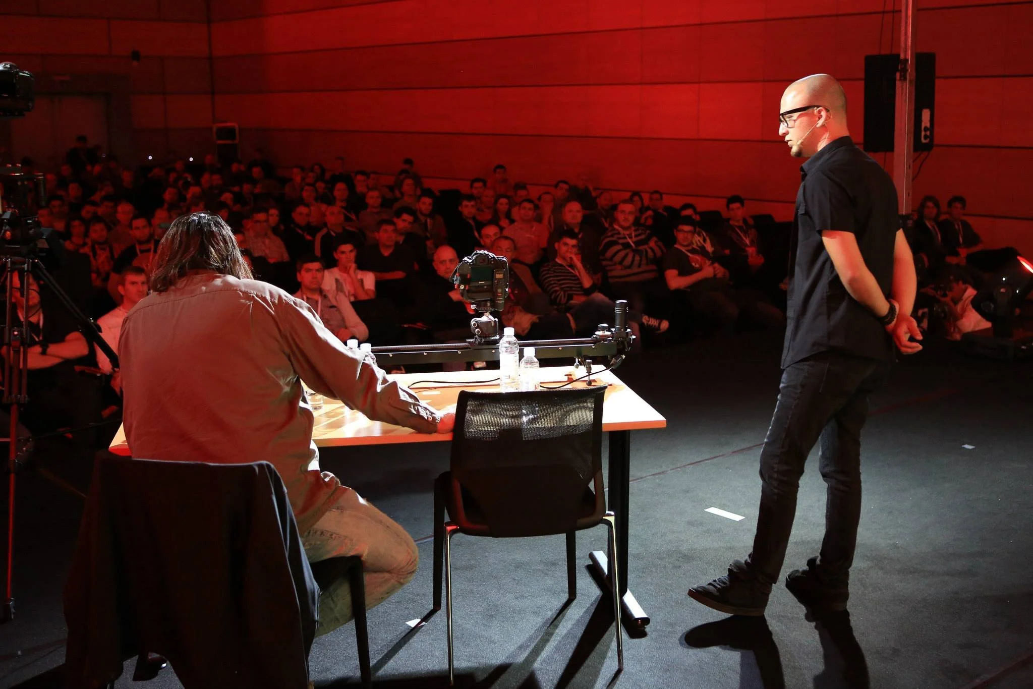 A man with glasses and a bald head standing on stage during a presentation, facing a seated presenter who has long hair and is wearing a beige jacket, with an audience watching in the background.