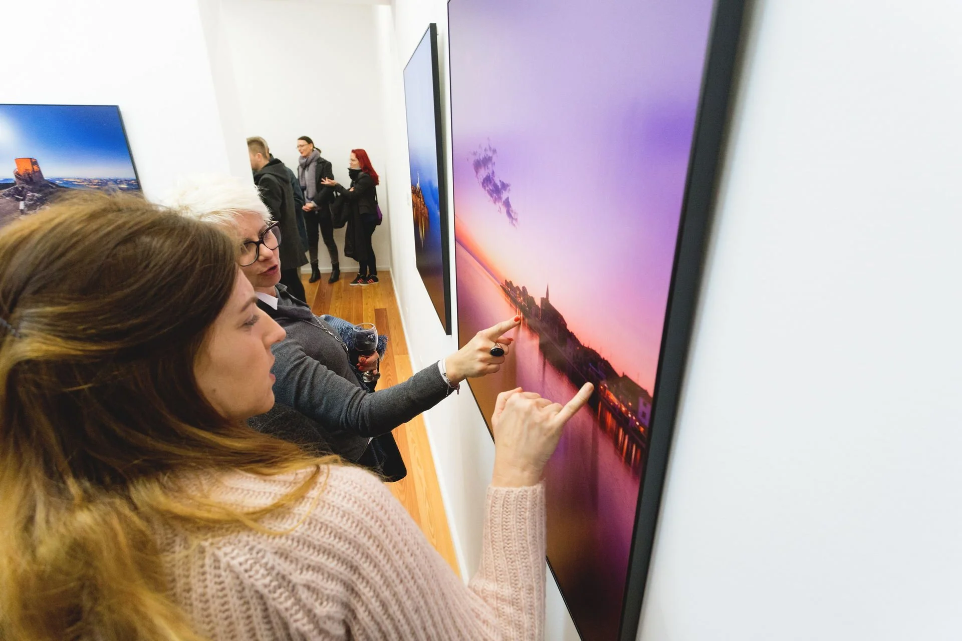 Two women are looking at a large flat-screen monitor displaying a sunset over a river with a church on a small island. One woman is pointing at the screen while the other is holding a glass of red wine. There are more people in the background observi