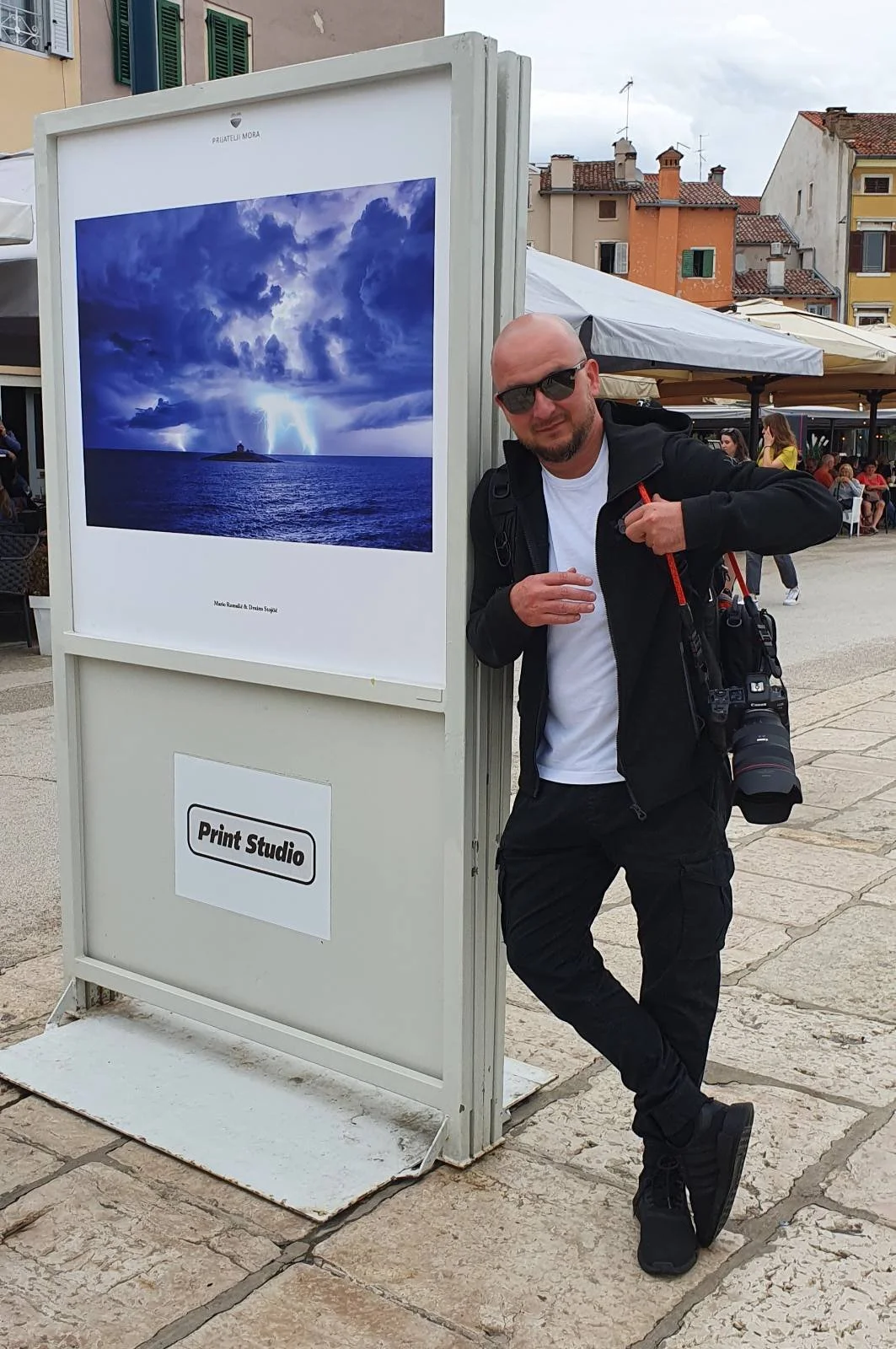 A man with sunglasses and a camera around his neck stands next to a large white display board featuring a photograph of a stormy sea with lightning and dramatic clouds over an island. The display board has a sign that says 'Print Studio.' The scene i