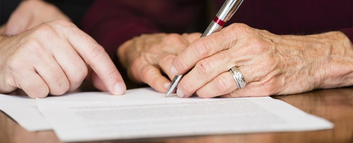 Lähikuva vanhemman henkilön käsistä allekirjoittamassa paperia.