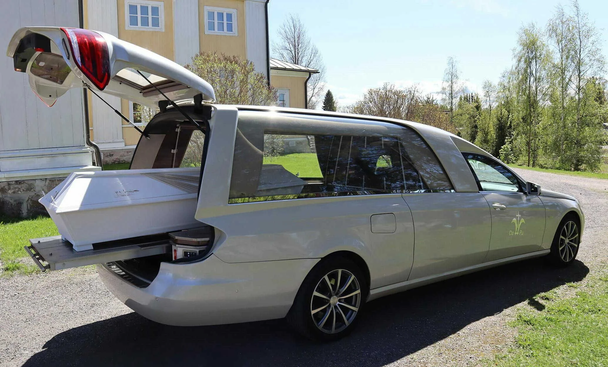 Kuljetusauto kirkon edustalla, valkoinen arkku osittain vielä auton sisällä.