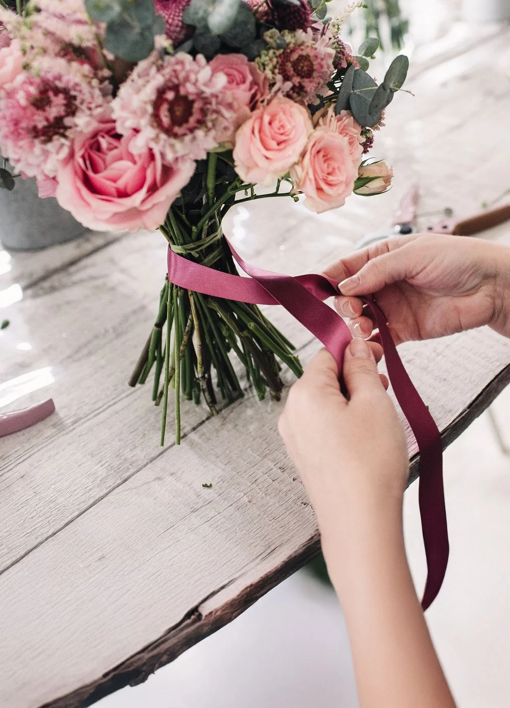 Lähikuva käsistä jotka solmivat vaaleanpunaista kukkakimppua.