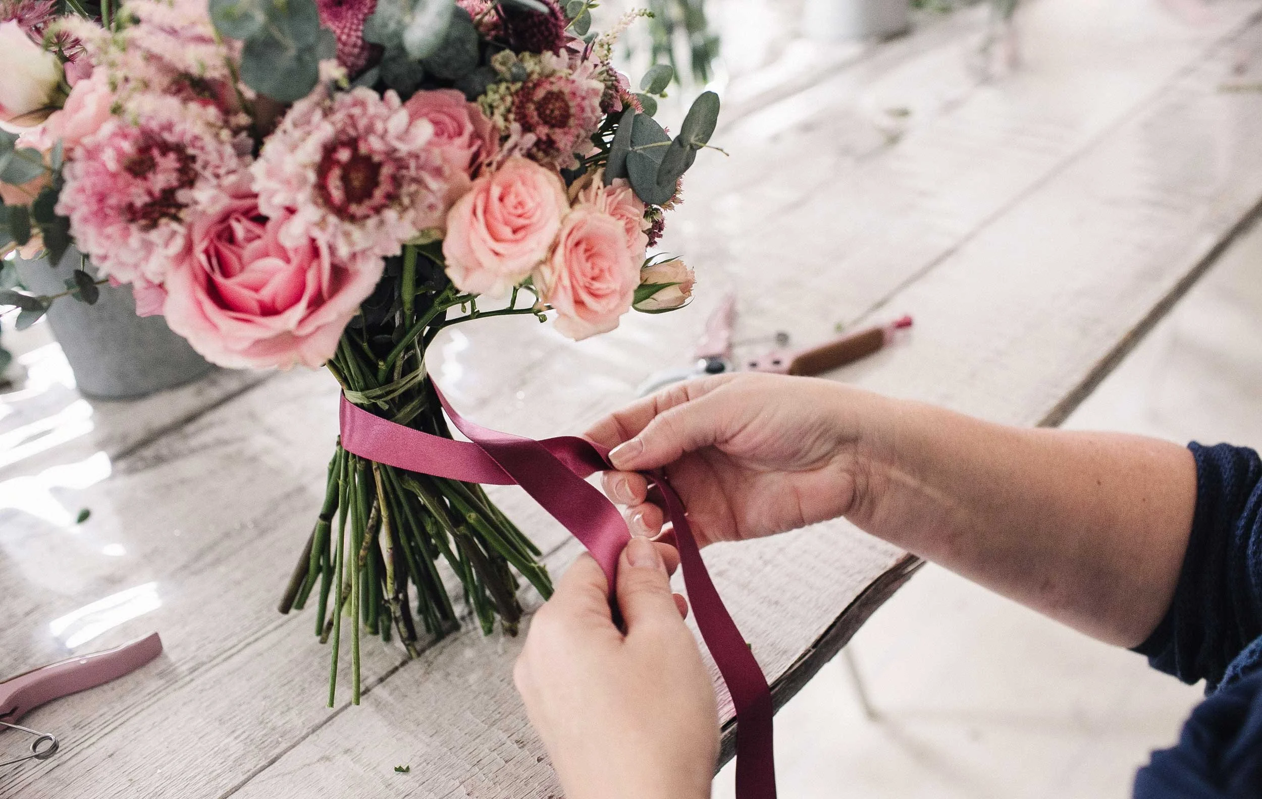 Lähikuva käsistä jotka solmivat vaaleanpunaista kukkakimppua.