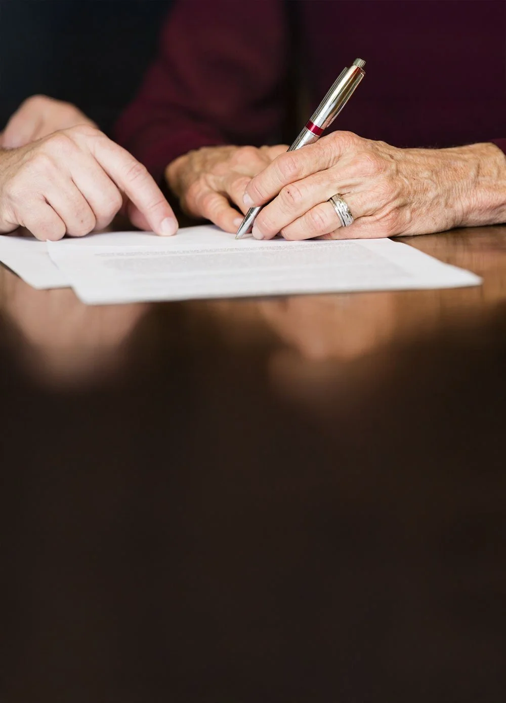 Lähikuva vanhemman henkilön käsistä, jotka allekirjoittavat paperia skeä vieressä toiset kädet näyttämässä jotain paperilta.