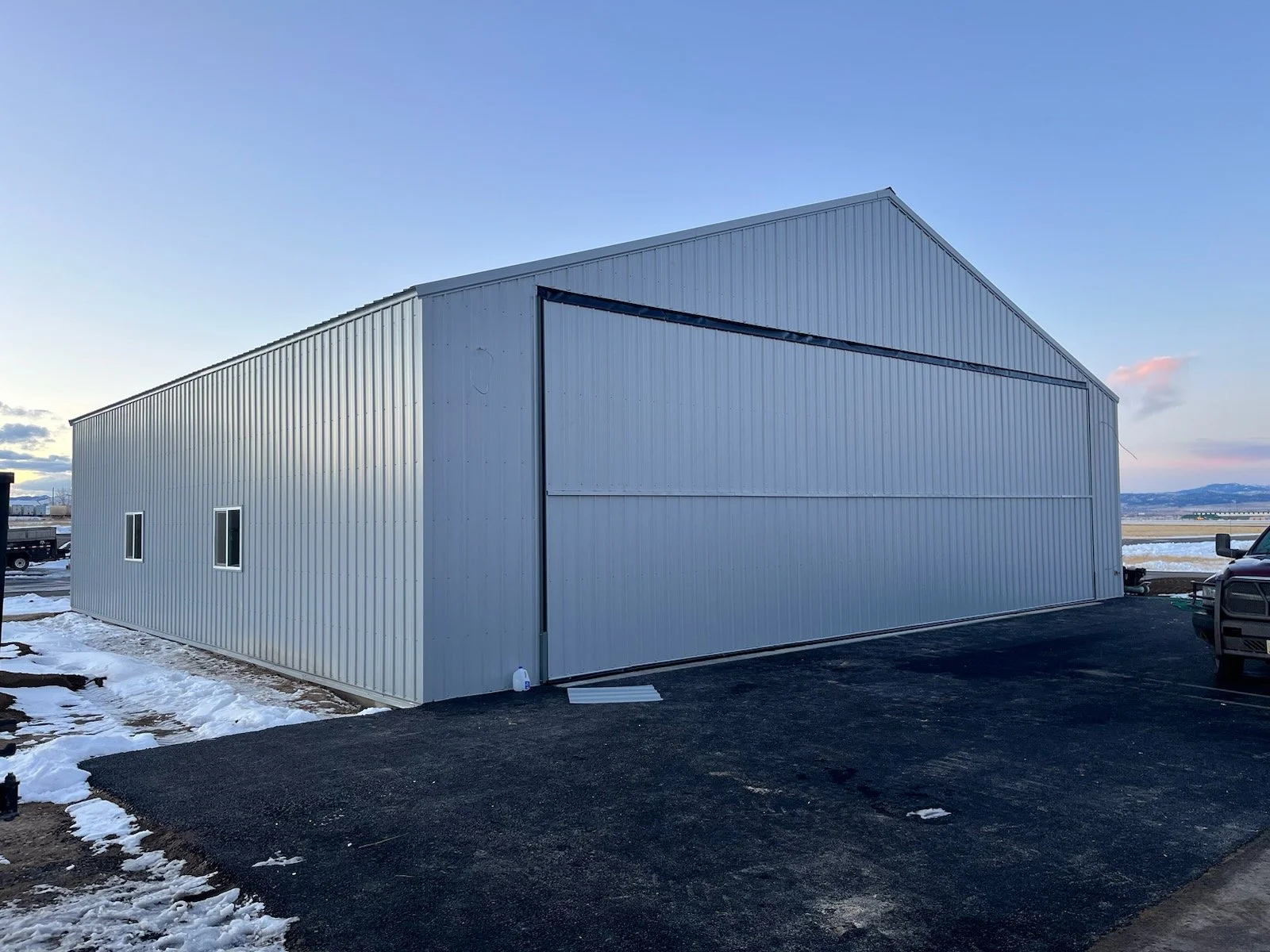 A large, white metal storage building with three small windows, situated on a paved surface with some snow around it, and a truck parked nearby on the right.