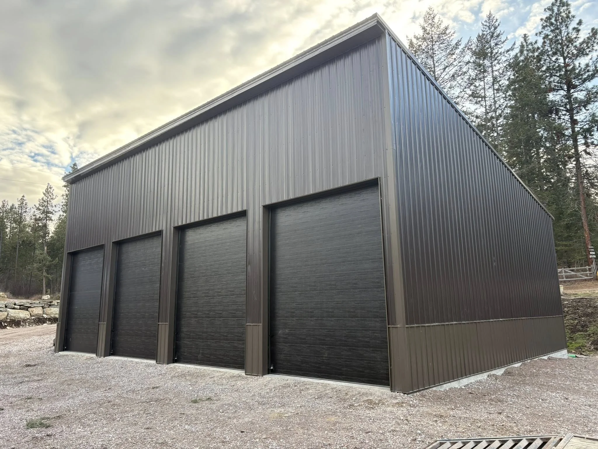 A large metal building with three black garage doors, set in a gravel area with trees in the background and a cloudy sky overhead.