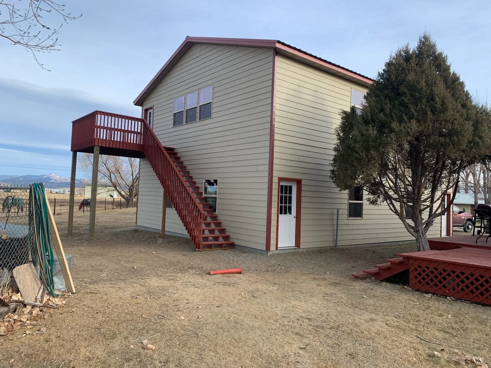 A two-story house with cream-colored siding, red trim, and a red staircase leading to a second-floor deck. There is a white door on the ground level, a window above it, and three windows on the upper floor. A large tree is on the right side of the house, and the yard is mostly dirt with some sparse grass.