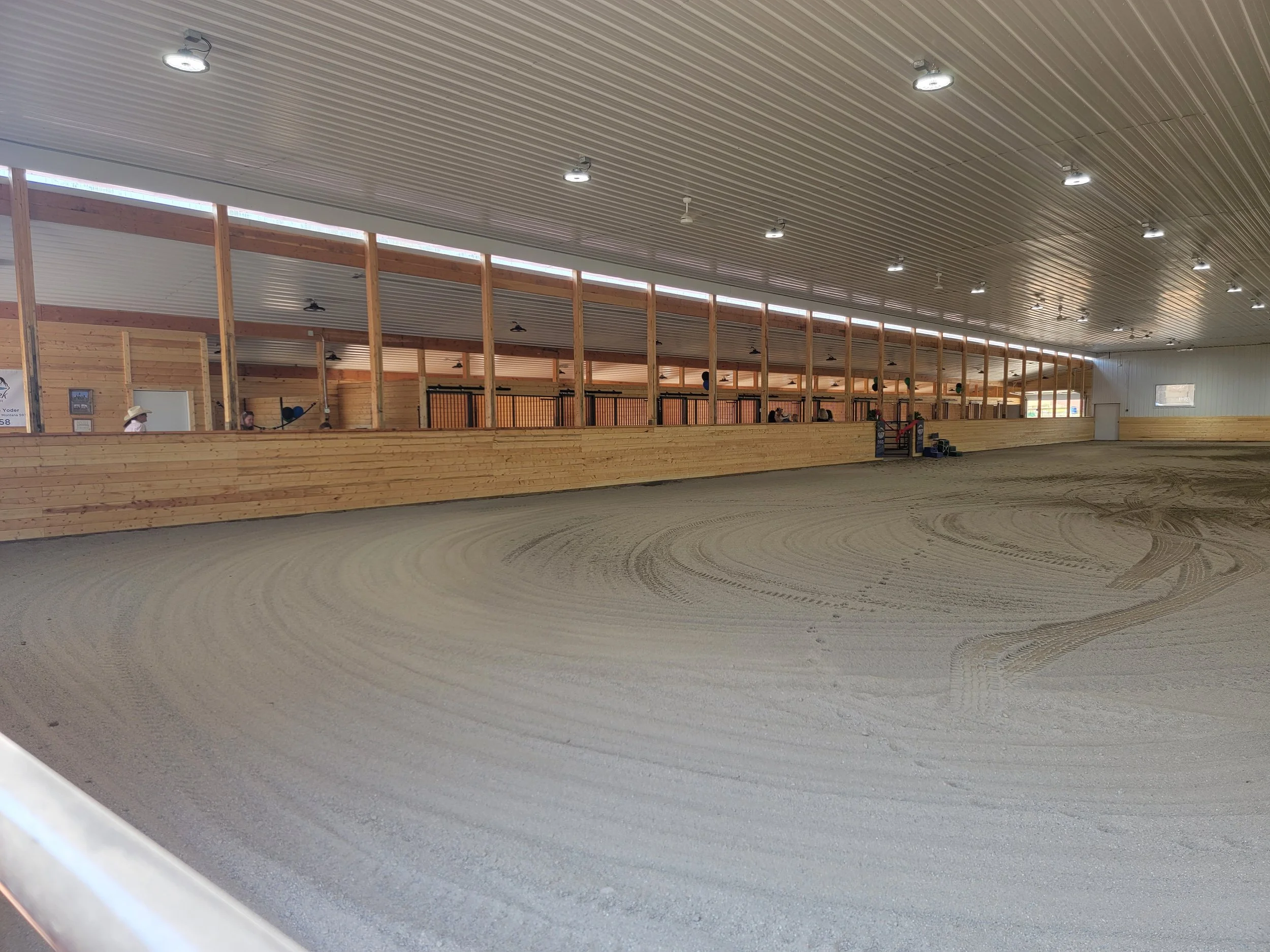 Indoor horse riding arena with a sandy track, wooden railings, and seating area with people