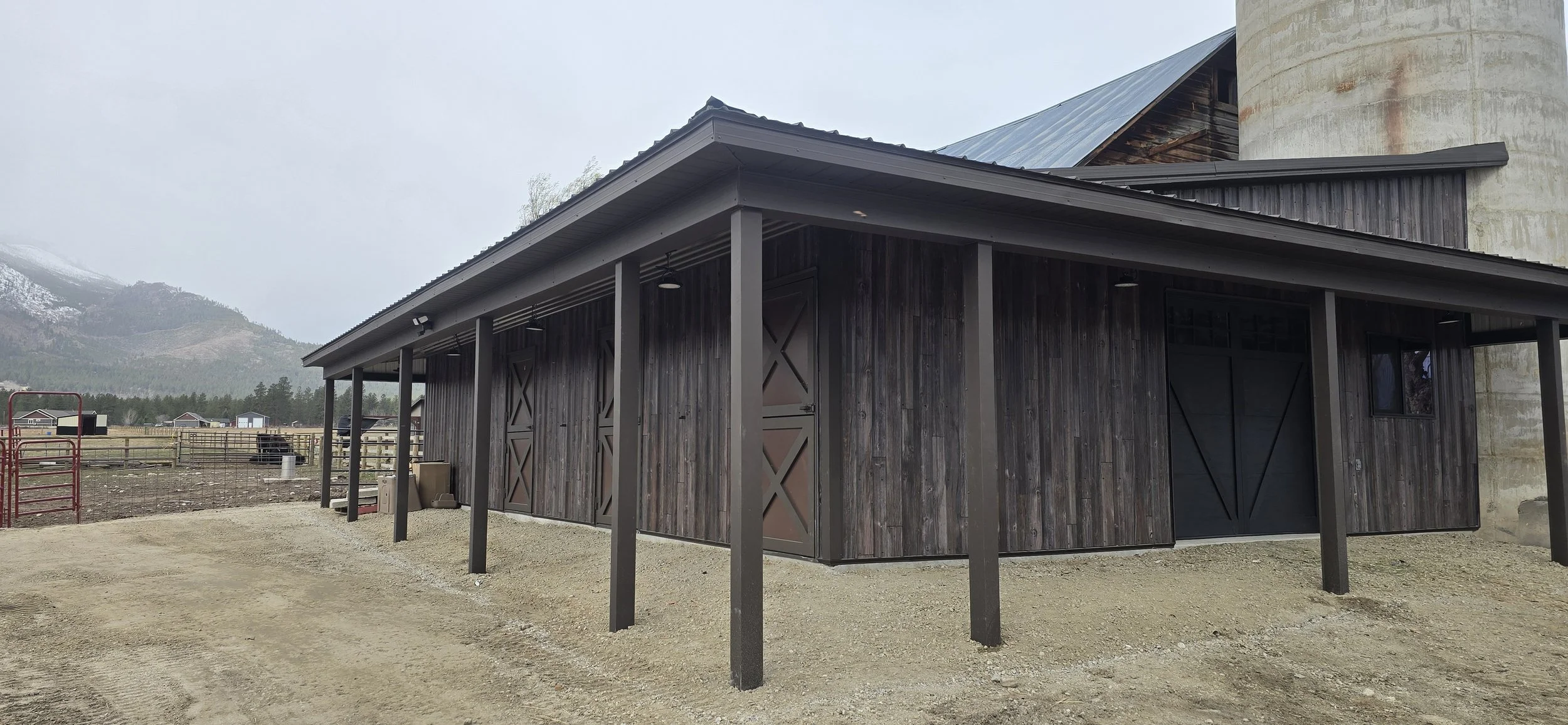 Construction of a wooden and metal shed outdoors with plywood walls and a metal roof, in a rural area with mountains in the background, a dirt ground in the foreground, and a part of a yellow piece of equipment to the right.