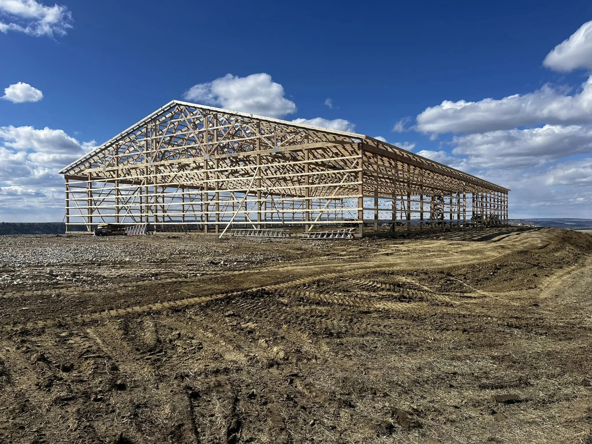 Wooden framework of a large building under construction outdoors on a clear day with blue sky and scattered clouds, on a dirt and gravel ground.