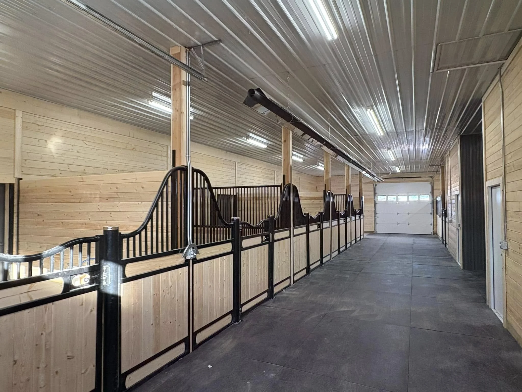 Indoor horse stable with wooden walls and black metal stalls, concrete floor, and overhead lights. Large garage door at the far end.