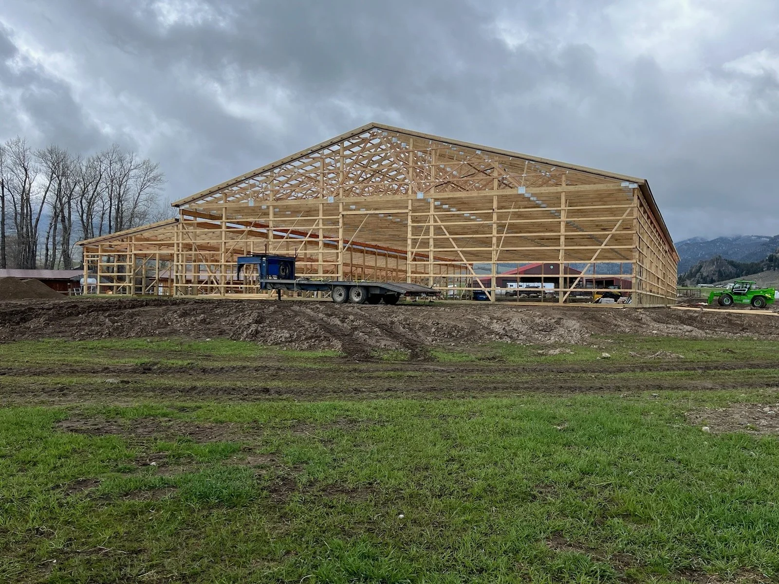 A building under construction with visible wooden framework, set on a grassy and dirt landscape, with vehicles and equipment on site, and mountains and cloudy sky in the background.