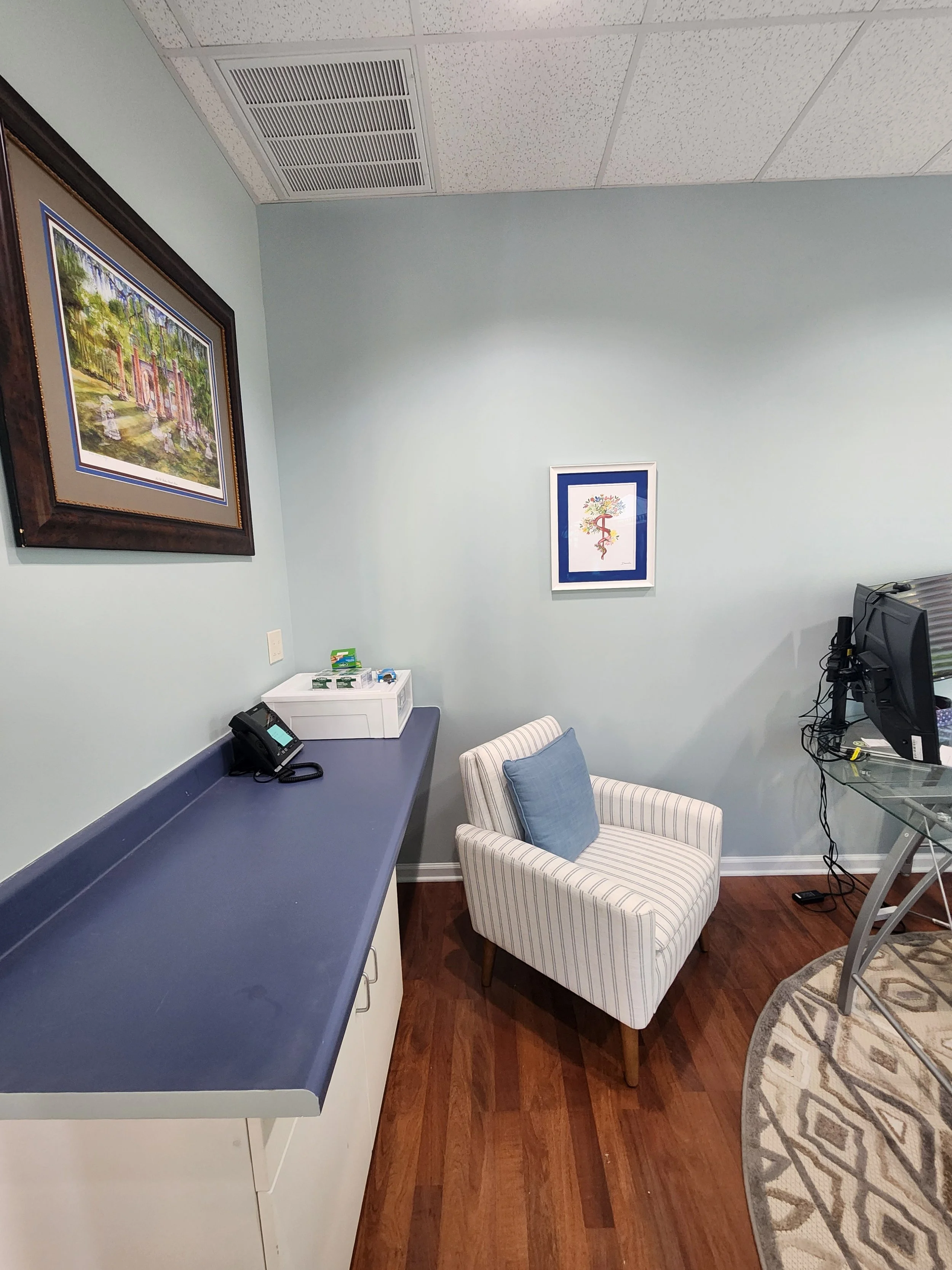 Office with a light blue wall, a striped armchair with a blue cushion, a dark blue counter with a telephone, and framed artwork on the walls, hardwood floor, and part of a glass desk with a computer.