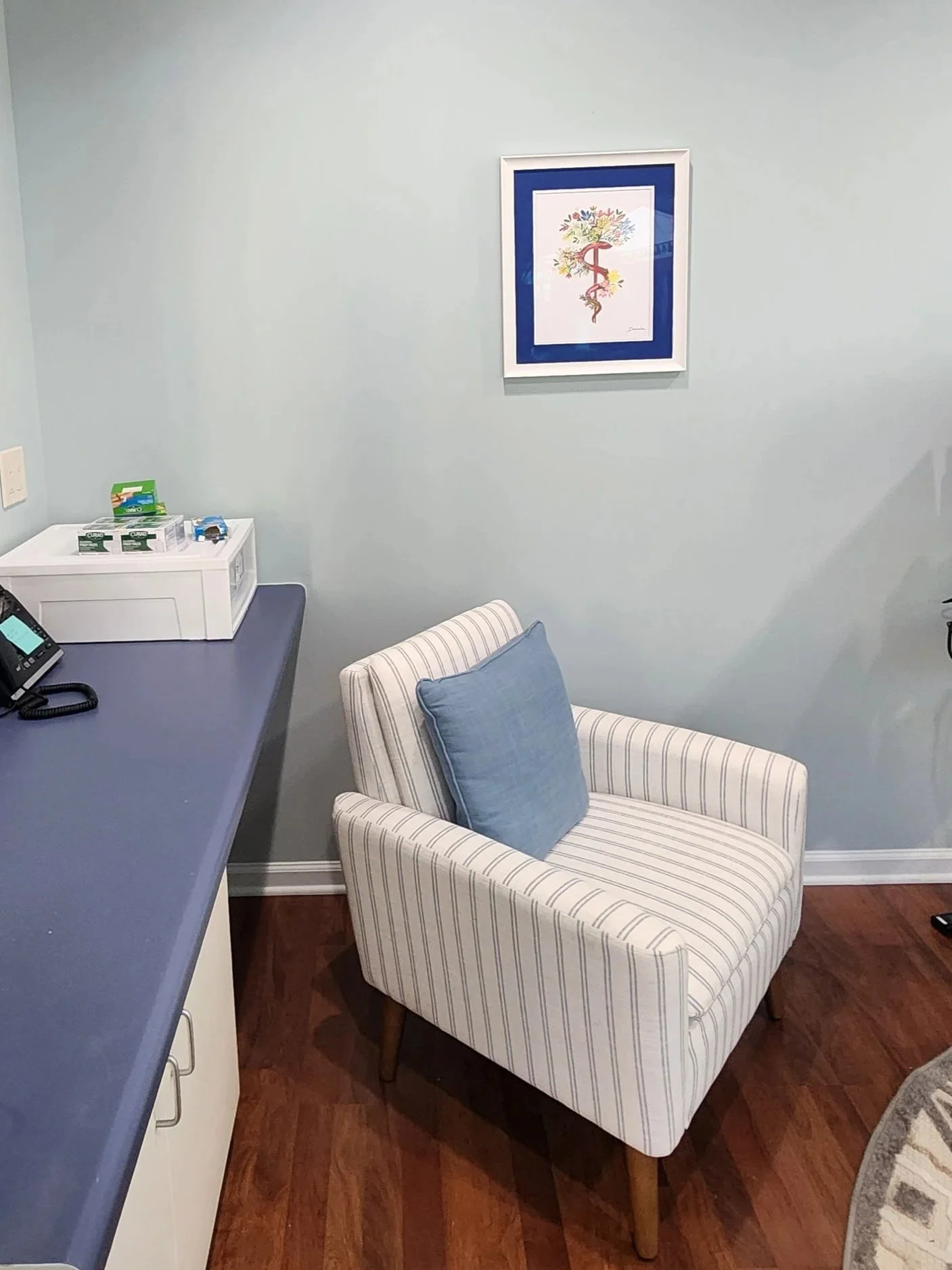 A waiting room with a striped armchair with a blue pillow, a wall artwork of a tree, a countertop with medical supplies, and a paint-colored wall.