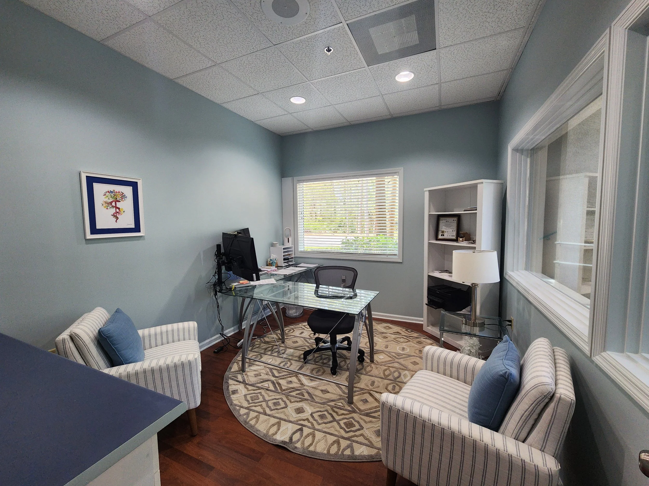 An office with two striped armchairs with blue pillows, a glass-top desk with a computer and office supplies, a white bookshelf, a small glass table with a lamp, a window with blinds, and a framed medical caduceus symbol on the wall.
