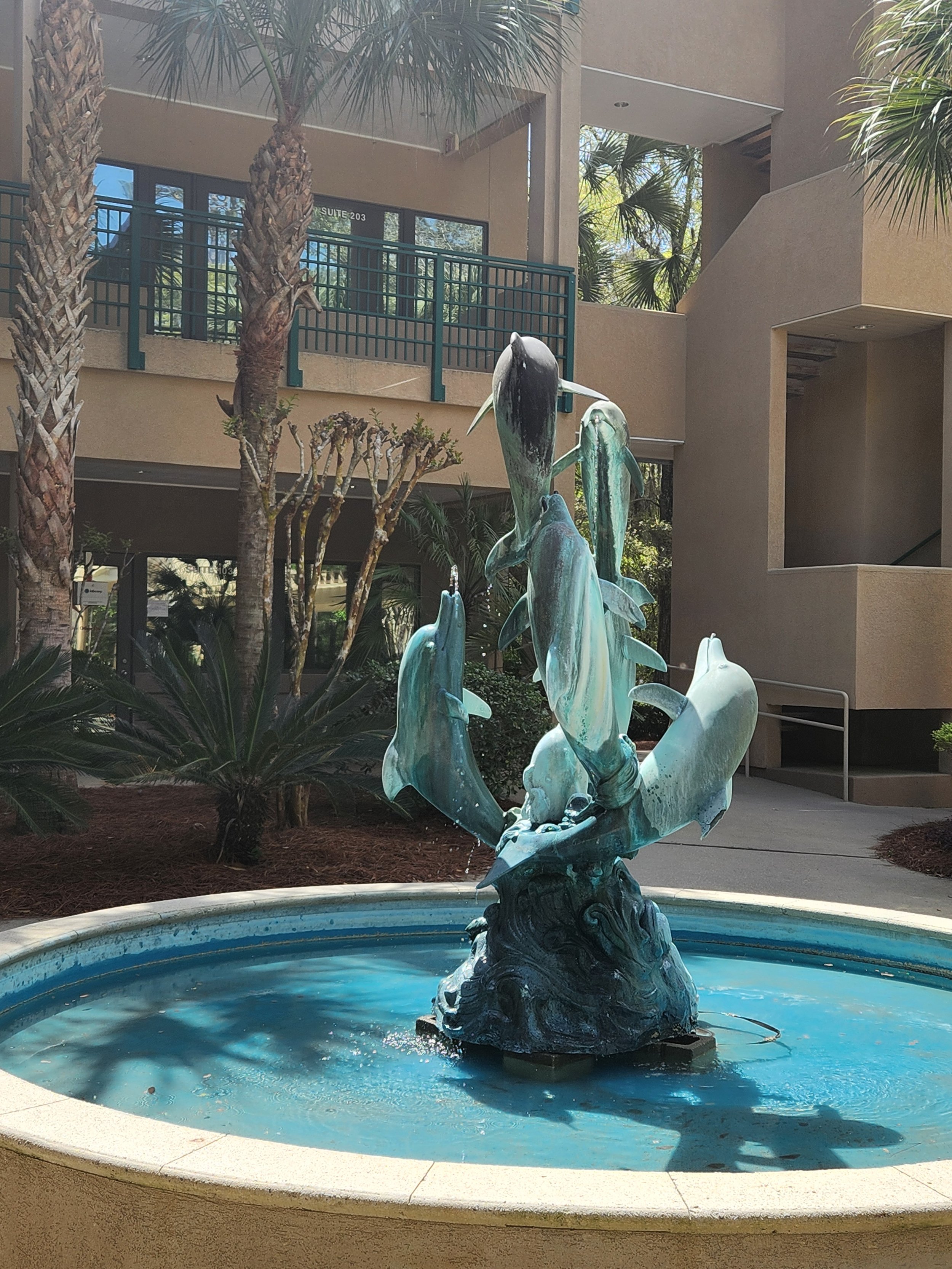 A bronze fountain sculpture of dolphins and a whale in a circular pool, located in an outdoor courtyard with palm trees and modern buildings.