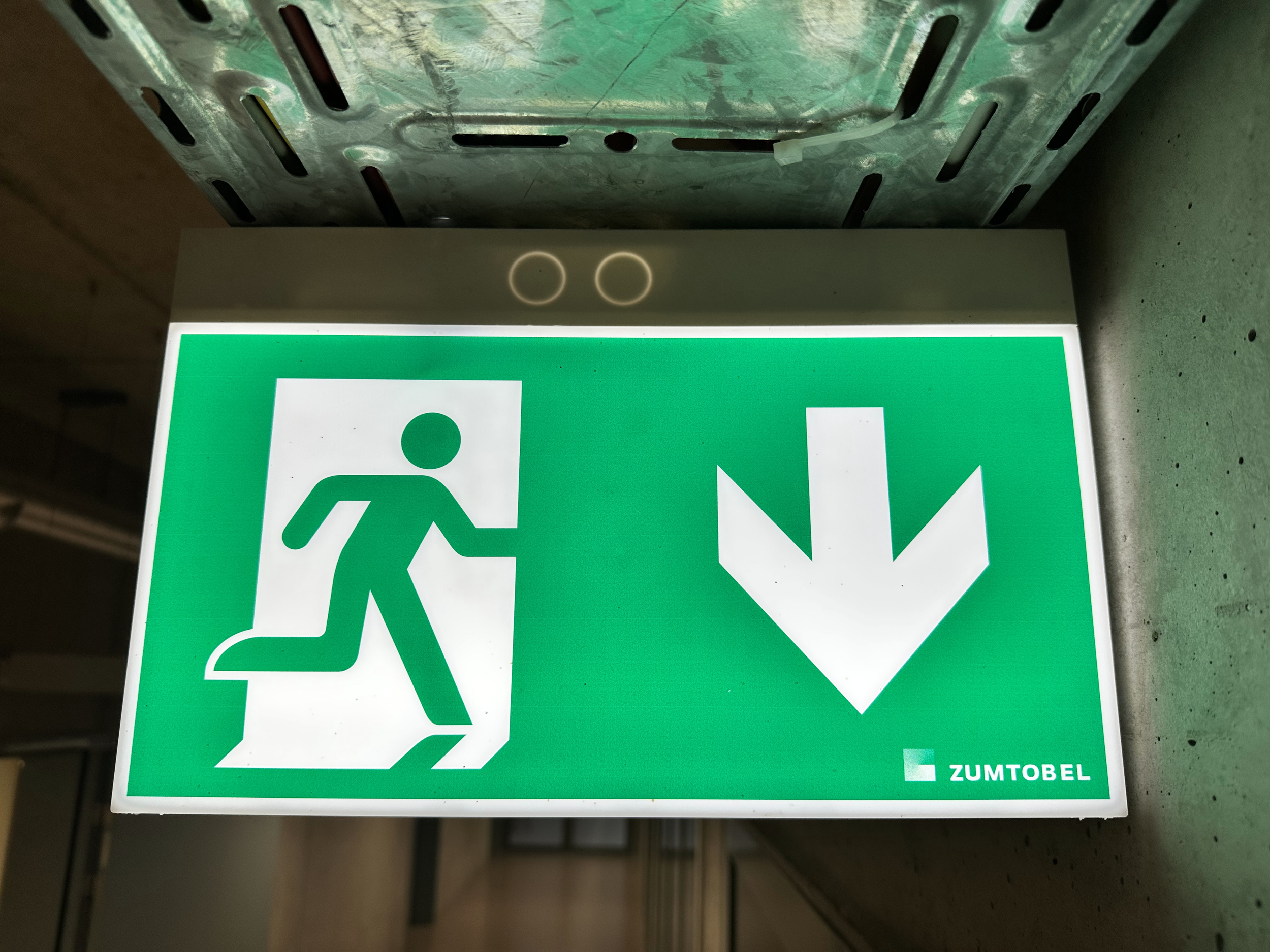 Grünes Notausgangsschild mit Fluchtwegpiktogramm und Pfeil nach unten, mit dem Text "ZUMTOBEL" am unteren rechten Rand.